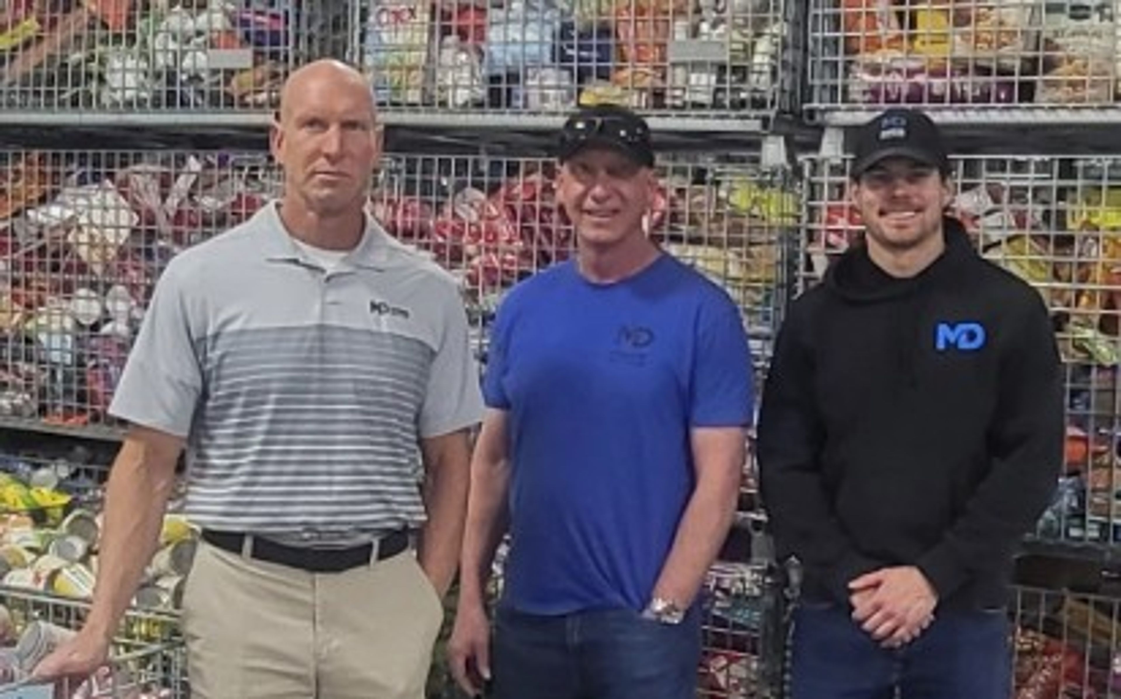 Three men stand in front of shelves filled with canned goods; two wear blue shirts, one in gray. All have “MD” logos on their shirts.