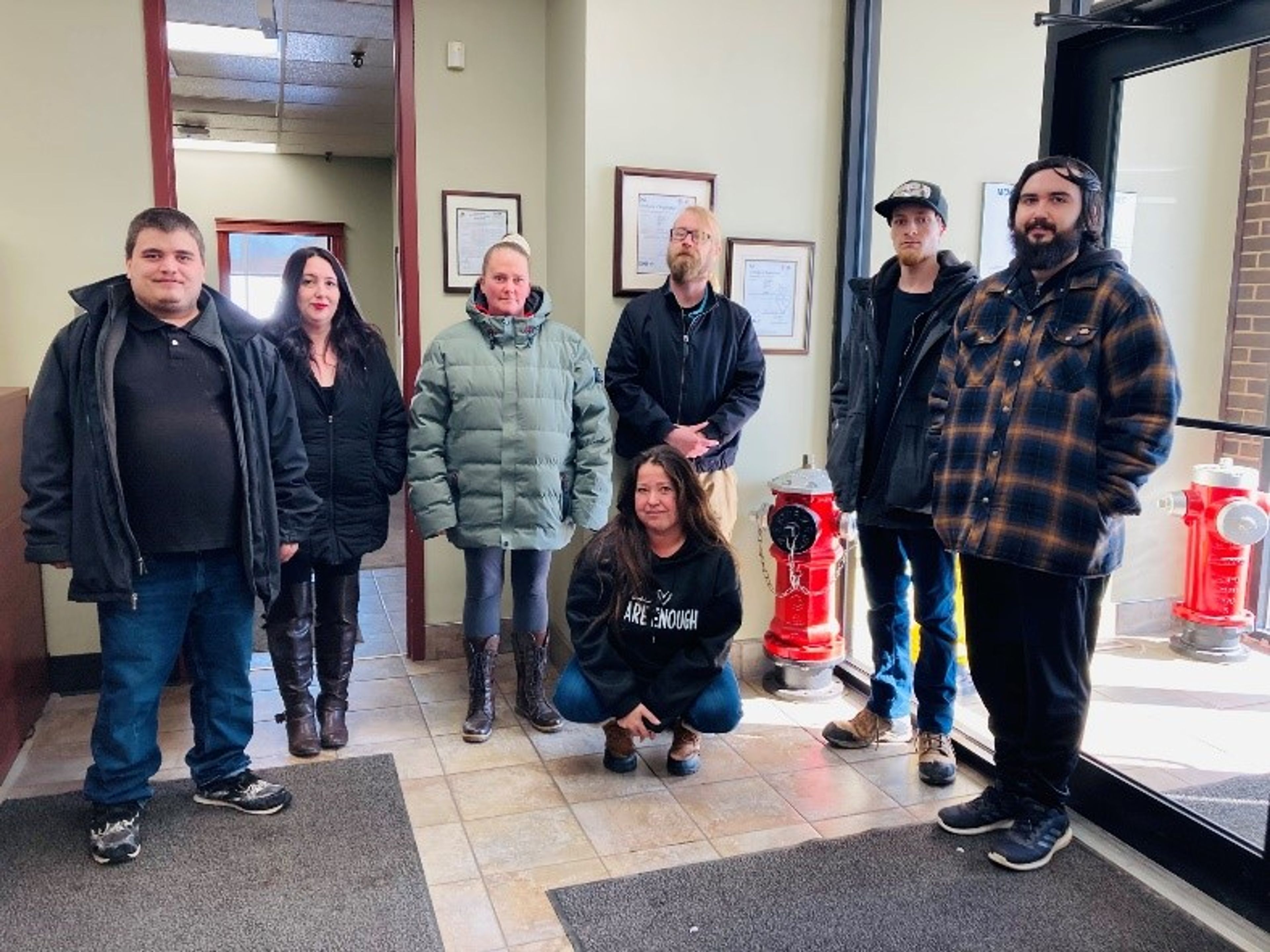 Seven people standing indoors near red fire hydrants and framed certificates on the wall, with natural light coming through a door on the right. One person is kneeling in front, while the others stand in a line behind her.