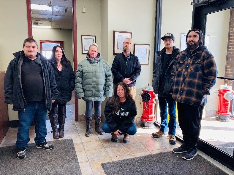 Seven people standing indoors near red fire hydrants and framed certificates on the wall, with natural light coming through a door on the right. One person is kneeling in front, while the others stand in a line behind her.