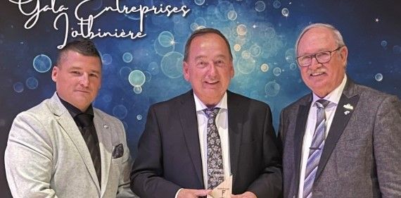 Three men in suits pose together at an event, one holding a small award. The background displays the text “Gala Entreprises Lotbinière” with a blue pattern and light spots.