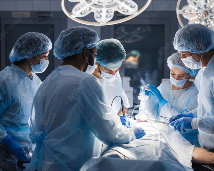 A group of surgeons in scrubs, masks, and gloves perform an operation under bright surgical lights in an operating room.