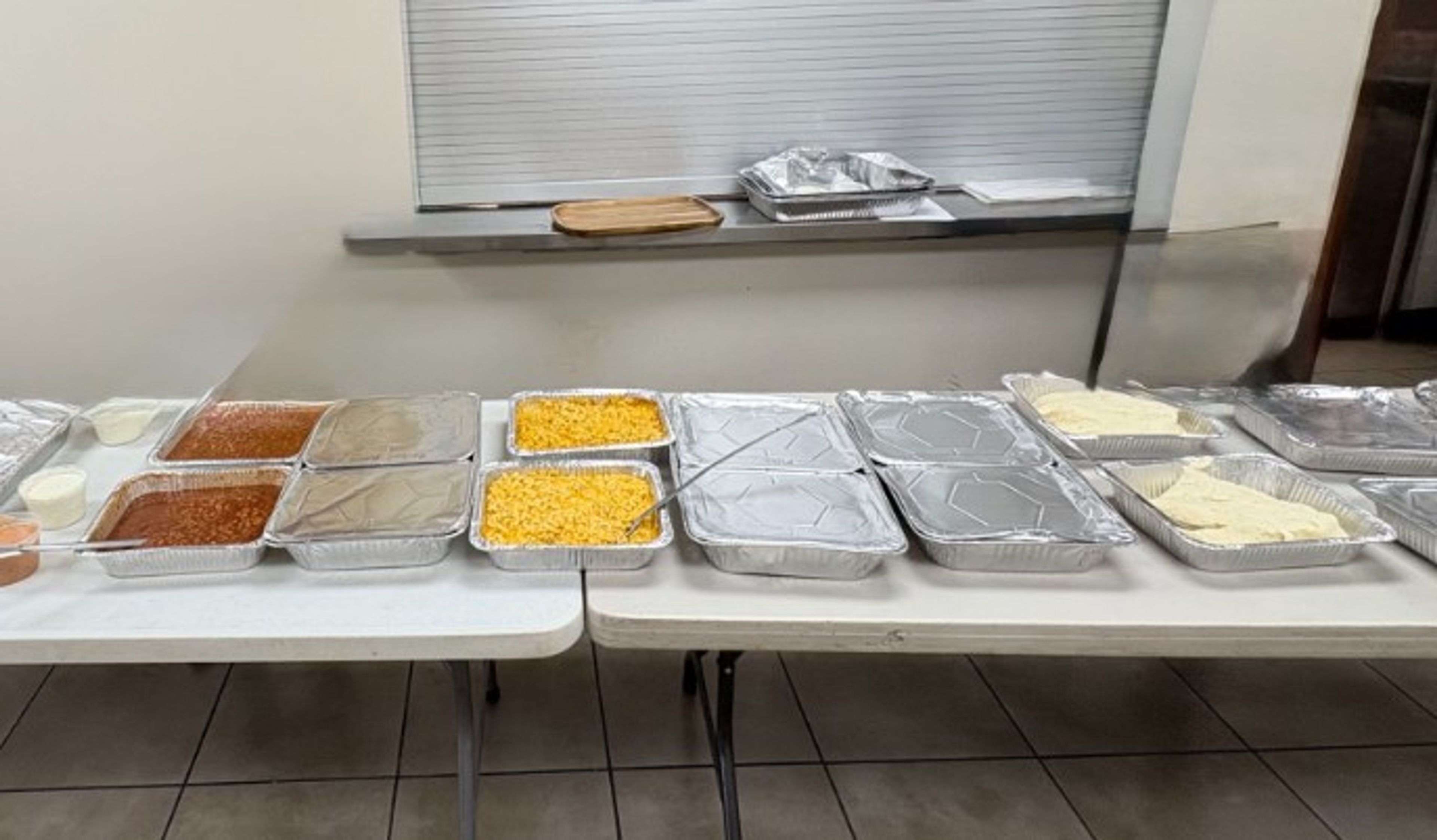 Several trays of food, some covered and some open, are arranged on white tables against a wall, with dishes including beans, corn, and what appears to be cheese or sauce.