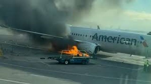 A baggage cart on fire near an American Airlines plane on the tarmac, with thick black smoke rising into the air.