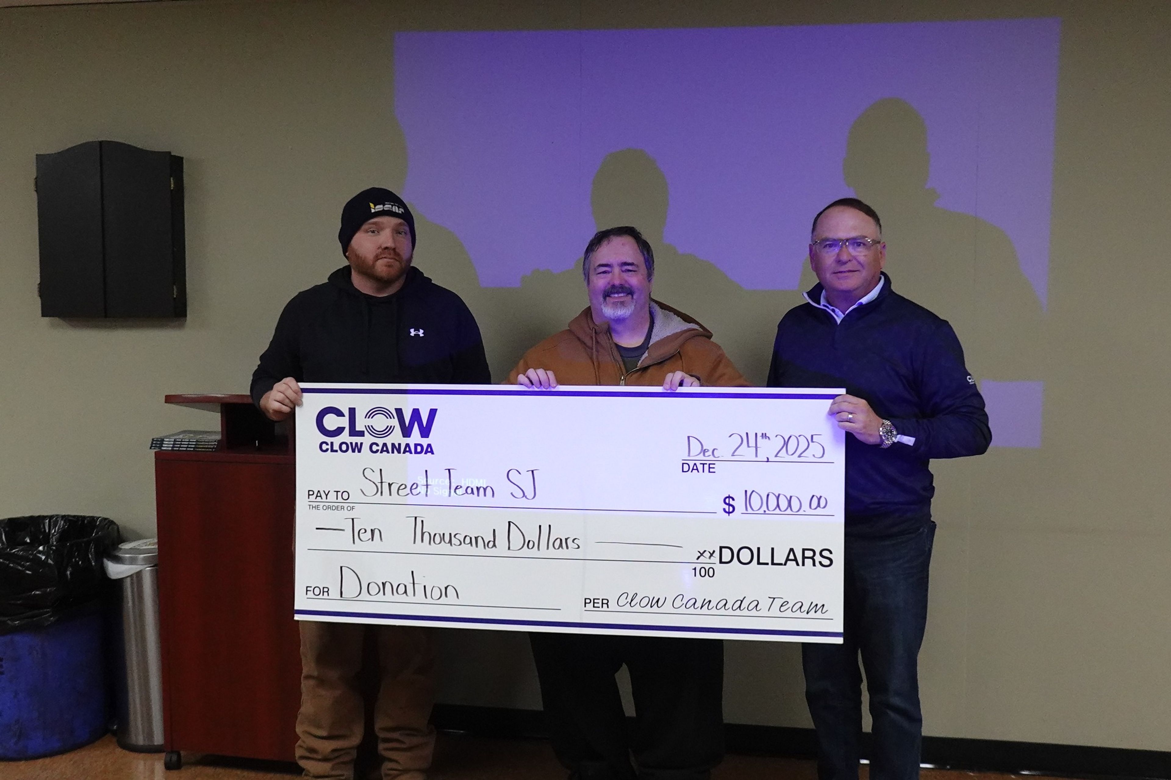 Three men stand indoors holding a large donation check for $10,000 made out to Street Team SJ from Clow Canada Team, dated December 21st, 2025.