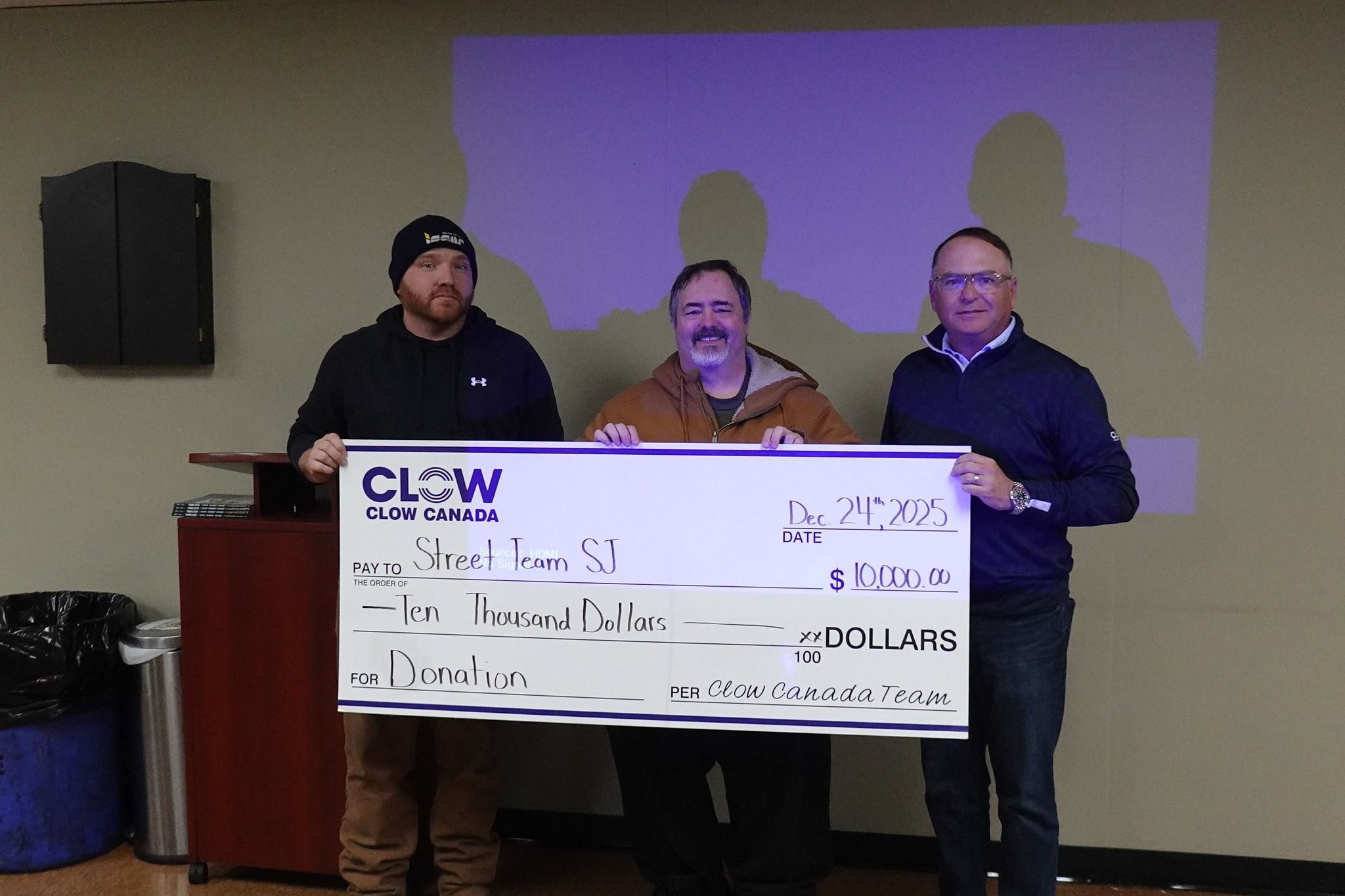 Three men stand indoors holding a large donation check for $10,000 made out to Street Team SJ from Clow Canada Team, dated December 21st, 2025.