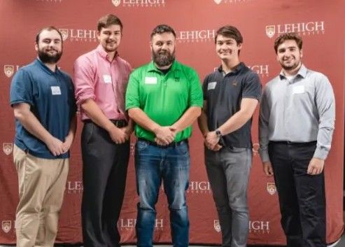 Five men stand in front of a Lehigh University backdrop, posing for a group photo. Four wear nametags and dress shirts, while the center man wears a green polo. All are smiling.