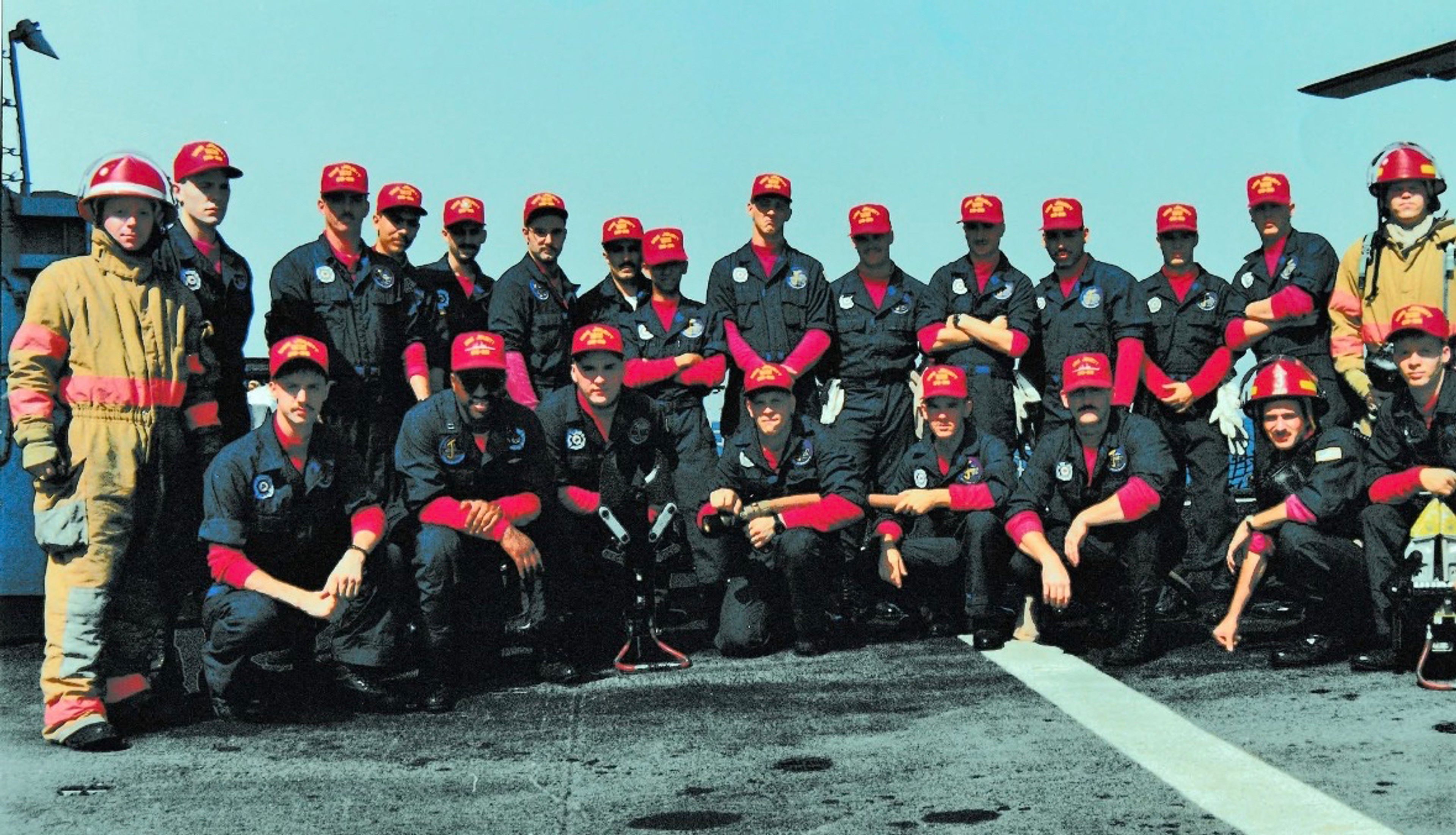 A group of firefighters in navy uniforms and red hats pose together outdoors on a concrete surface. Two members wear tan firefighting suits and helmets. It is sunny with a clear blue sky.