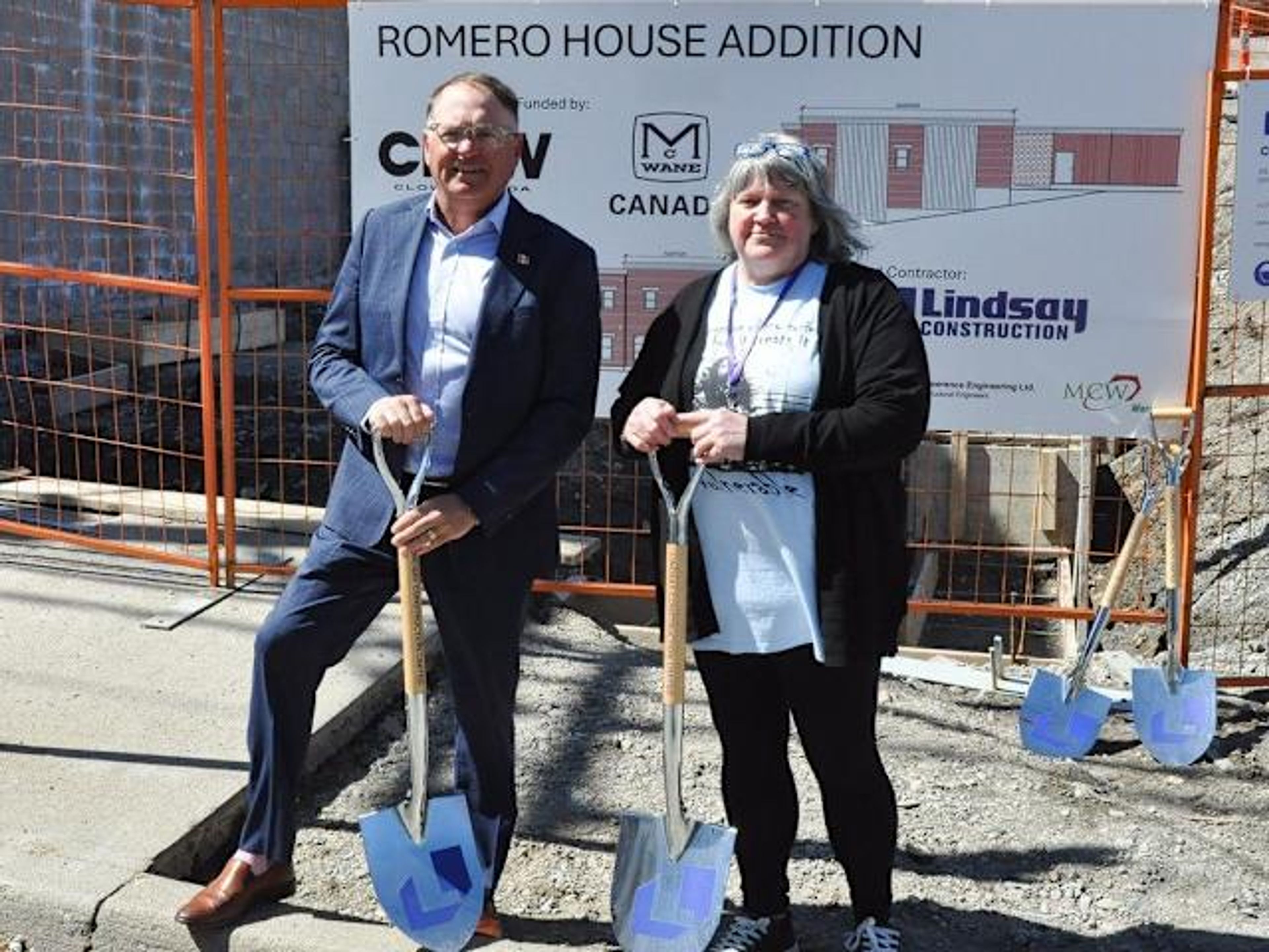A man in a suit and a woman in a cardigan hold shovels at a construction site, posing in front of a sign that reads “ROMERO HOUSE ADDITION.” They stand on a sidewalk next to an orange safety fence.