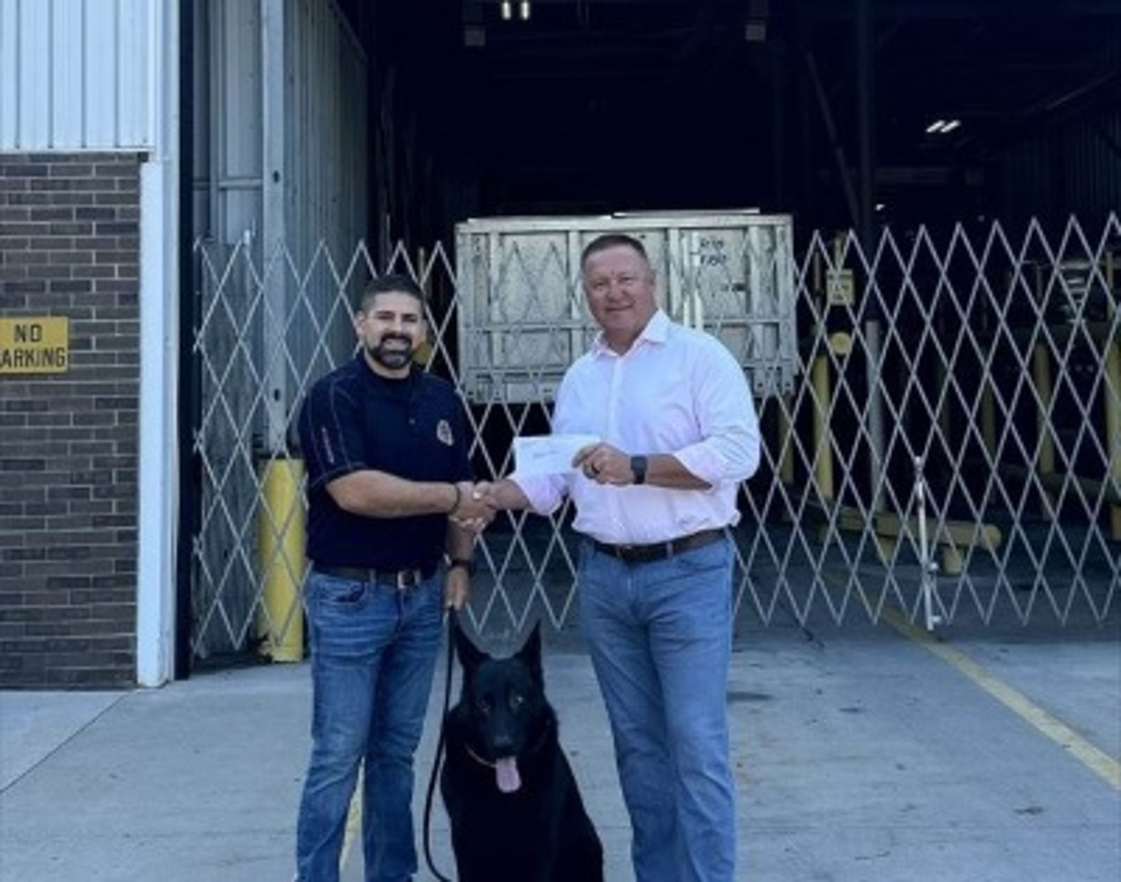 Two men shake hands while holding a piece of paper, standing outside an industrial building with a German Shepherd dog between them.