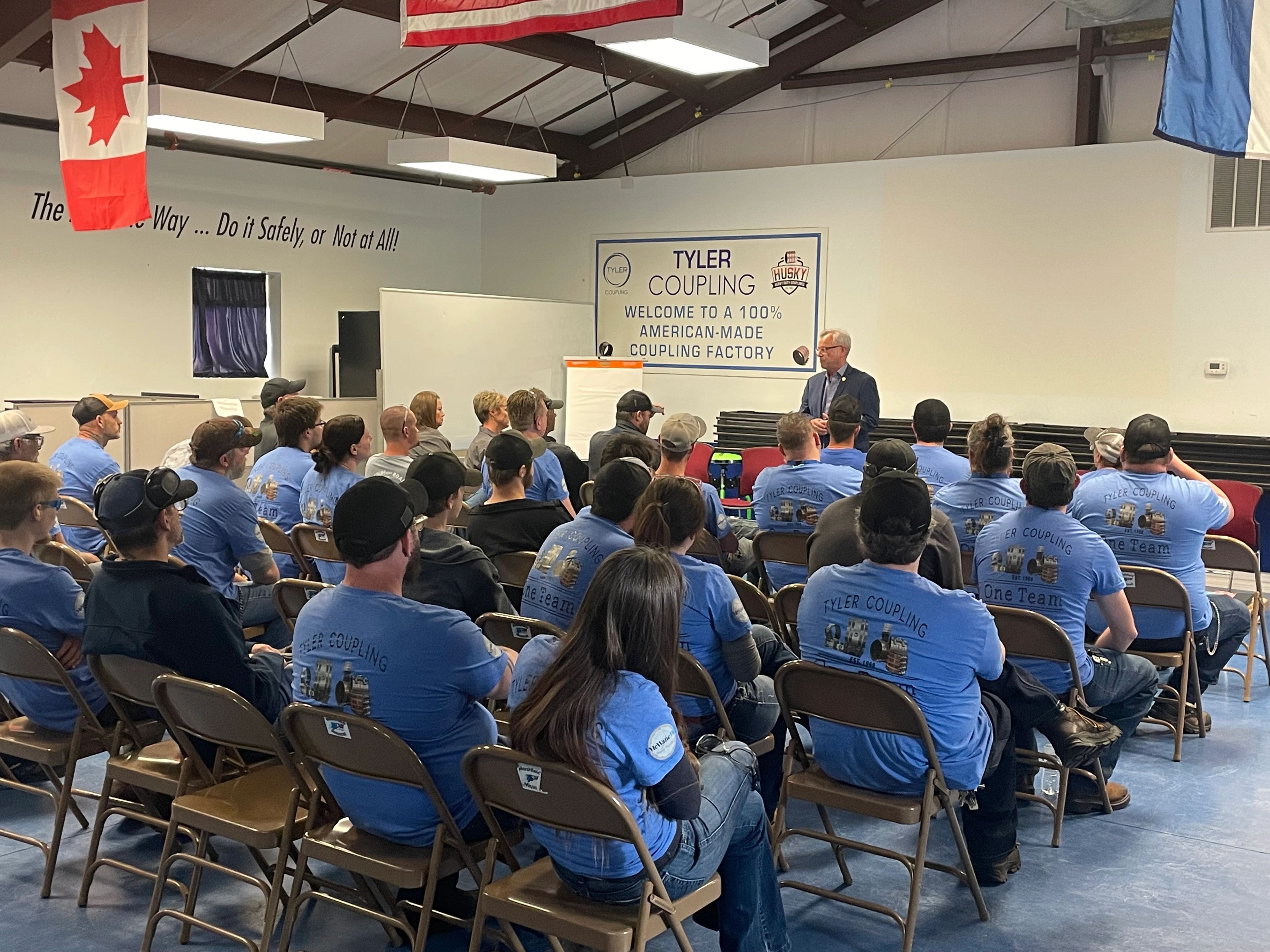 A group of people in matching blue shirts sit in folding chairs, listening to a speaker at the front of a room with banners and flags on the walls, including one that says “TYLER COUPLING.”.