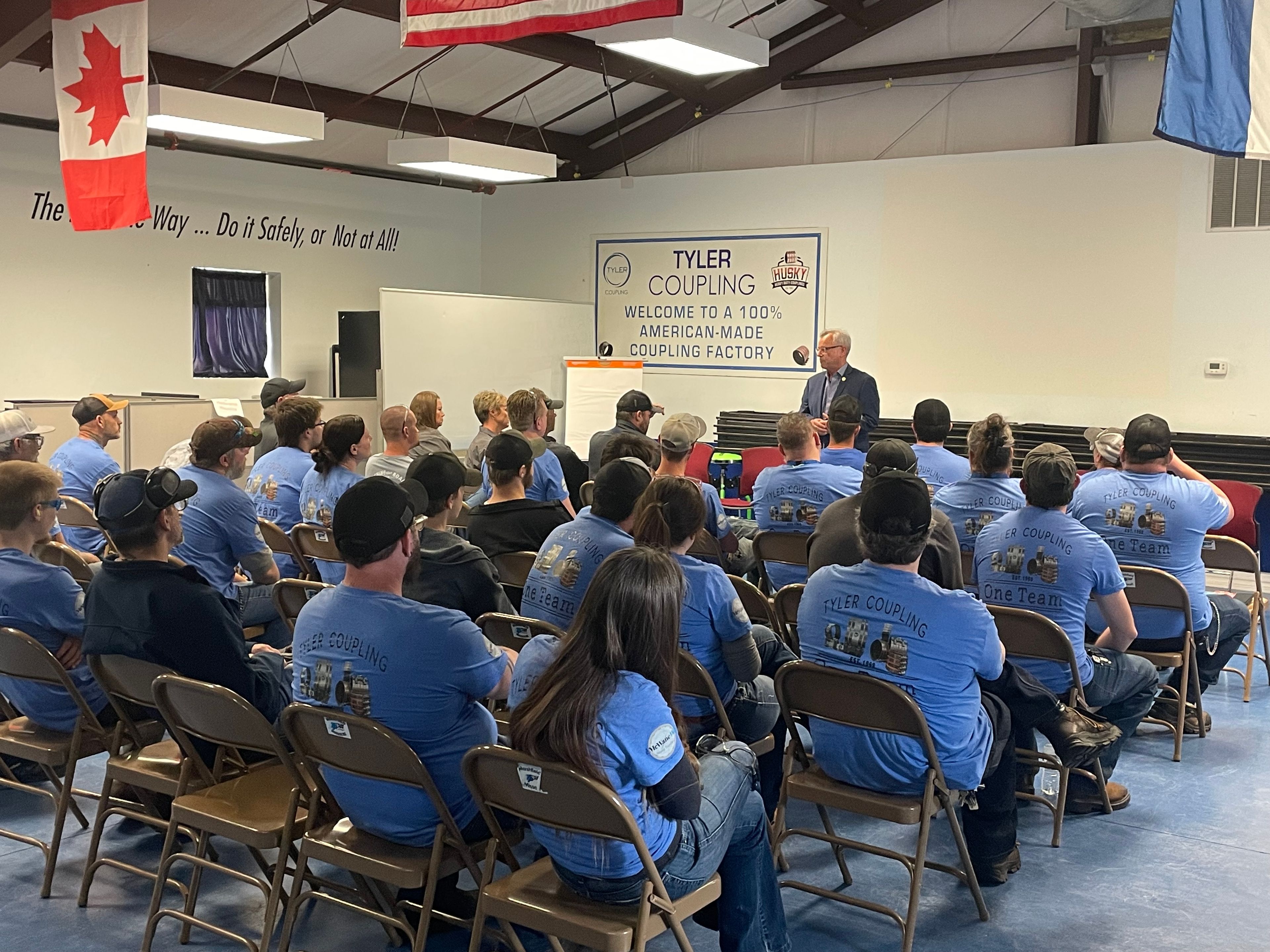 A group of people in matching blue shirts sit in folding chairs, listening to a speaker at the front of a room with banners and flags on the walls, including one that says “TYLER COUPLING.”.