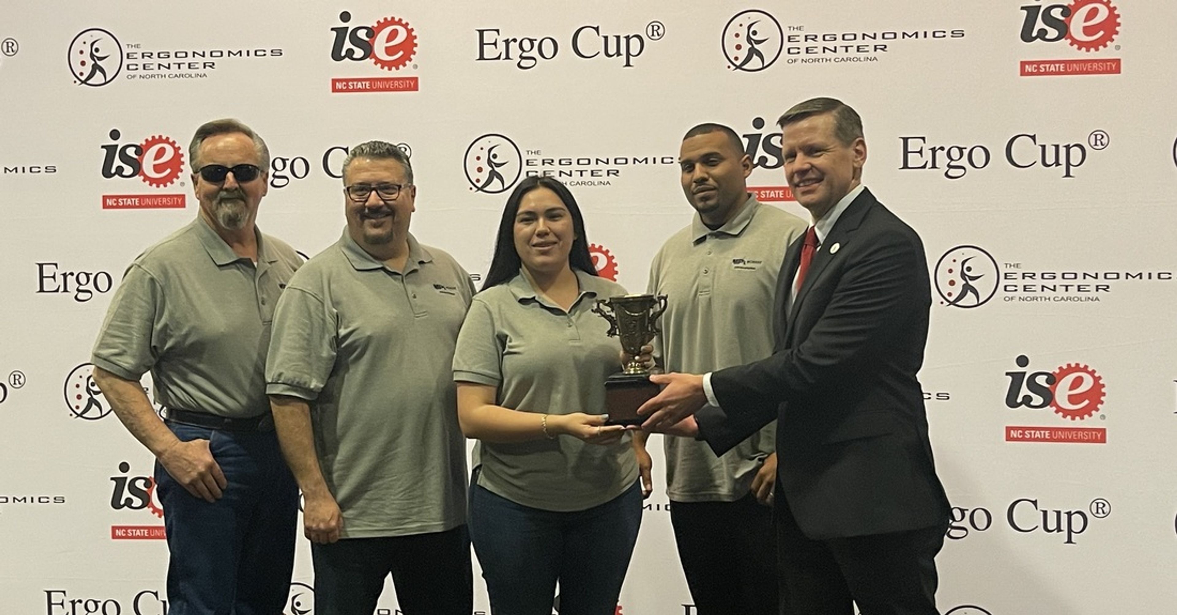 Four people in gray polos stand with a man in a suit, who is handing them a trophy. They pose in front of a backdrop with Ergo Cup and Ergonomics Center logos.
