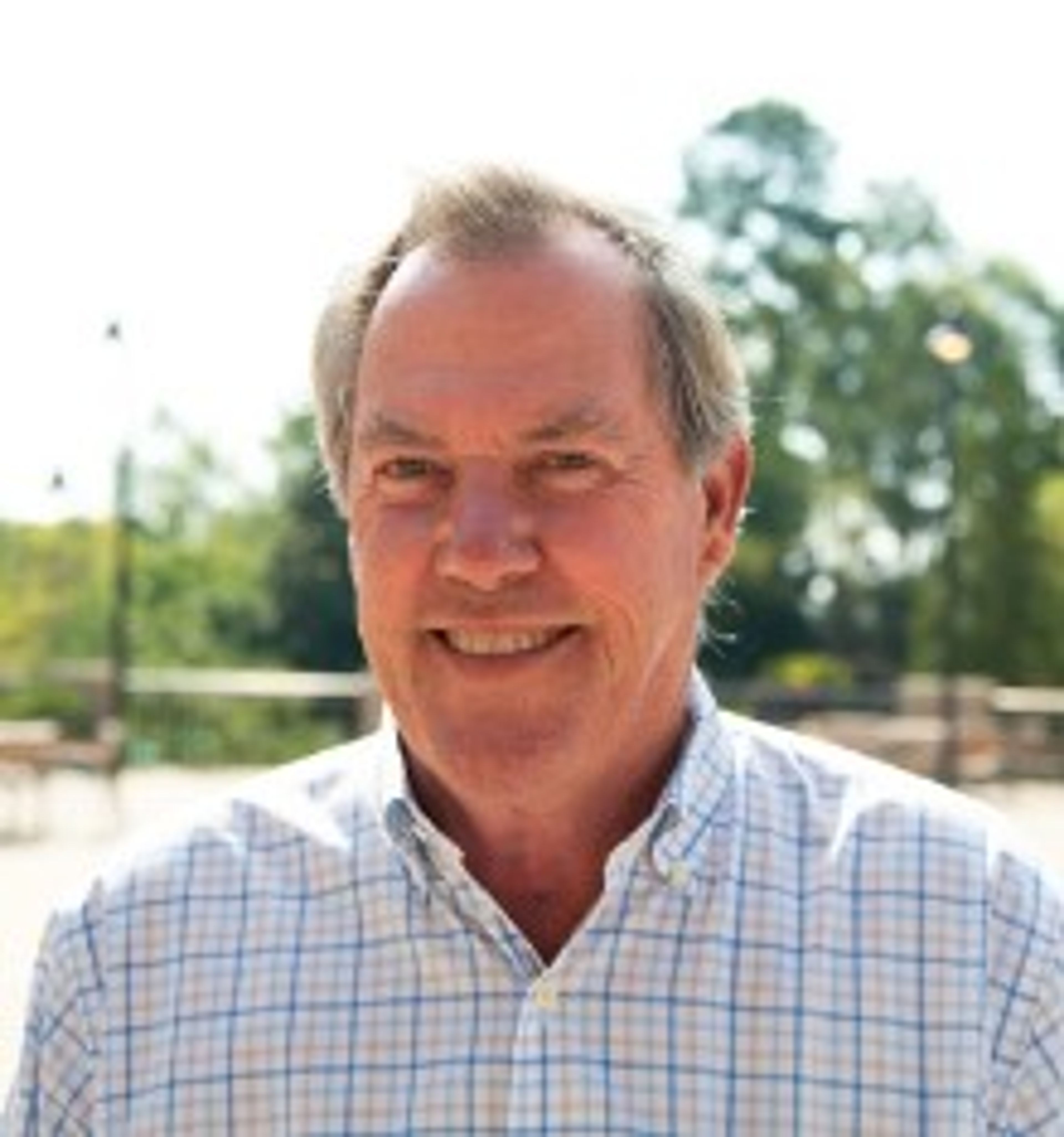 Smiling older man with gray hair wearing a light plaid shirt, standing outdoors with trees and a sunny background.