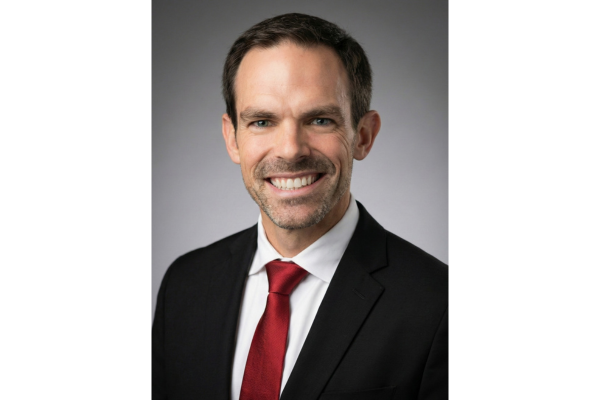 Smiling man in a black suit, white shirt, and red tie poses for a professional headshot against a plain gray background.