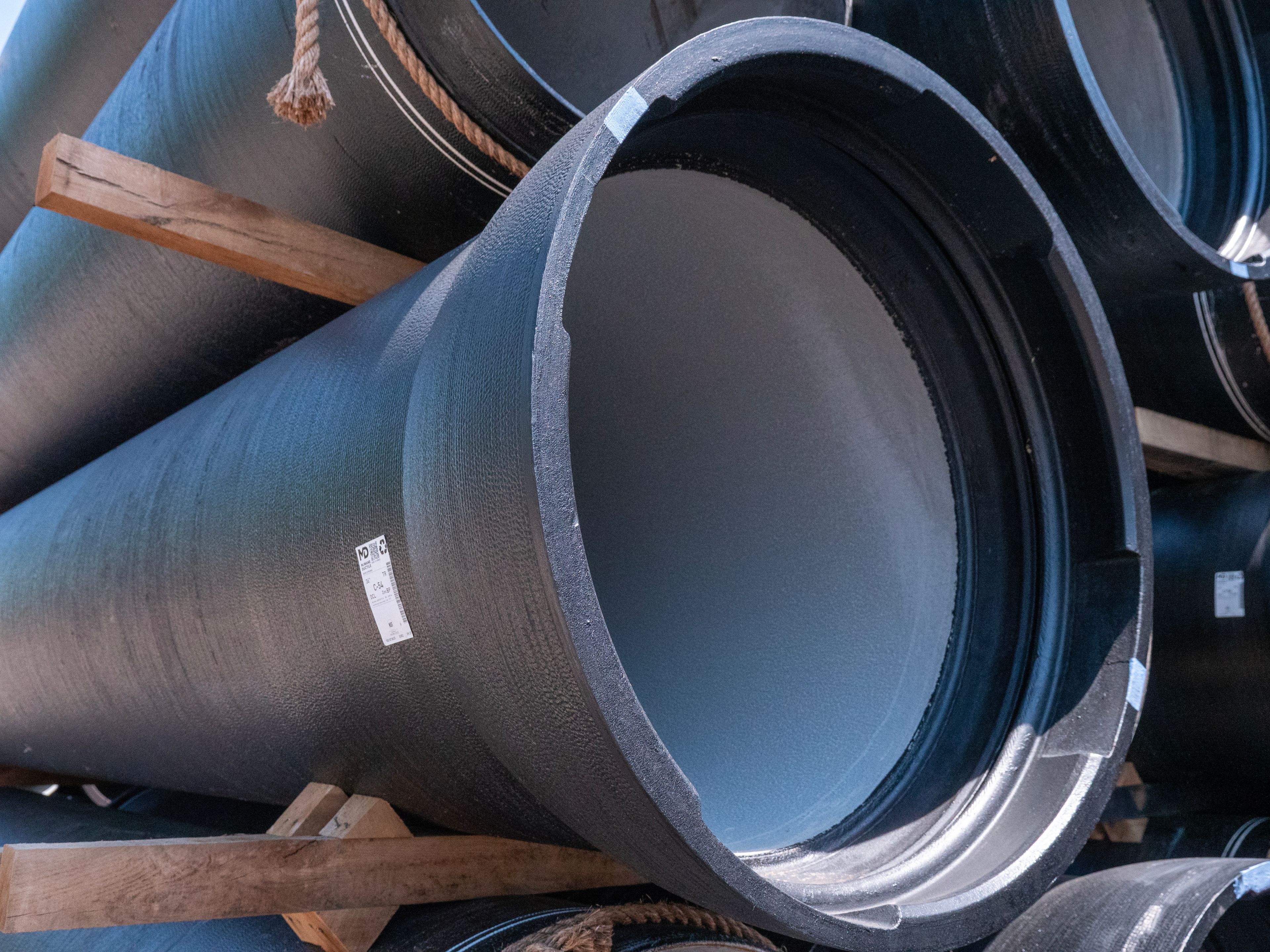 Close-up view of large black industrial pipes stacked on top of each other, with wooden supports and ropes holding them in place. The pipes have labels attached to their surfaces.