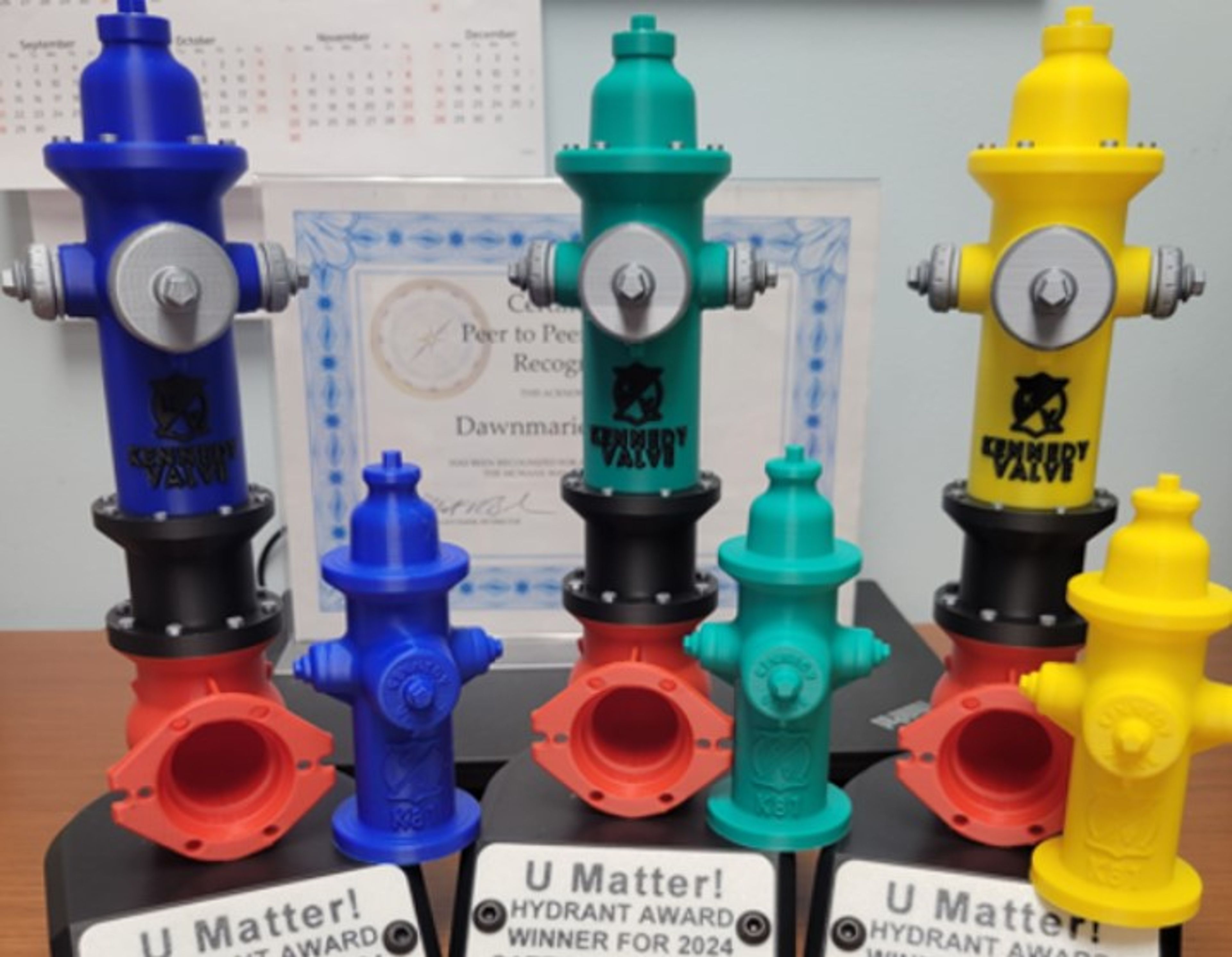 Colorful plastic fire hydrant models in blue, red, yellow, and teal are displayed on stands labeled “U Matter! Hydrant Award Winner for 2024.” A certificate and calendar are visible in the background.