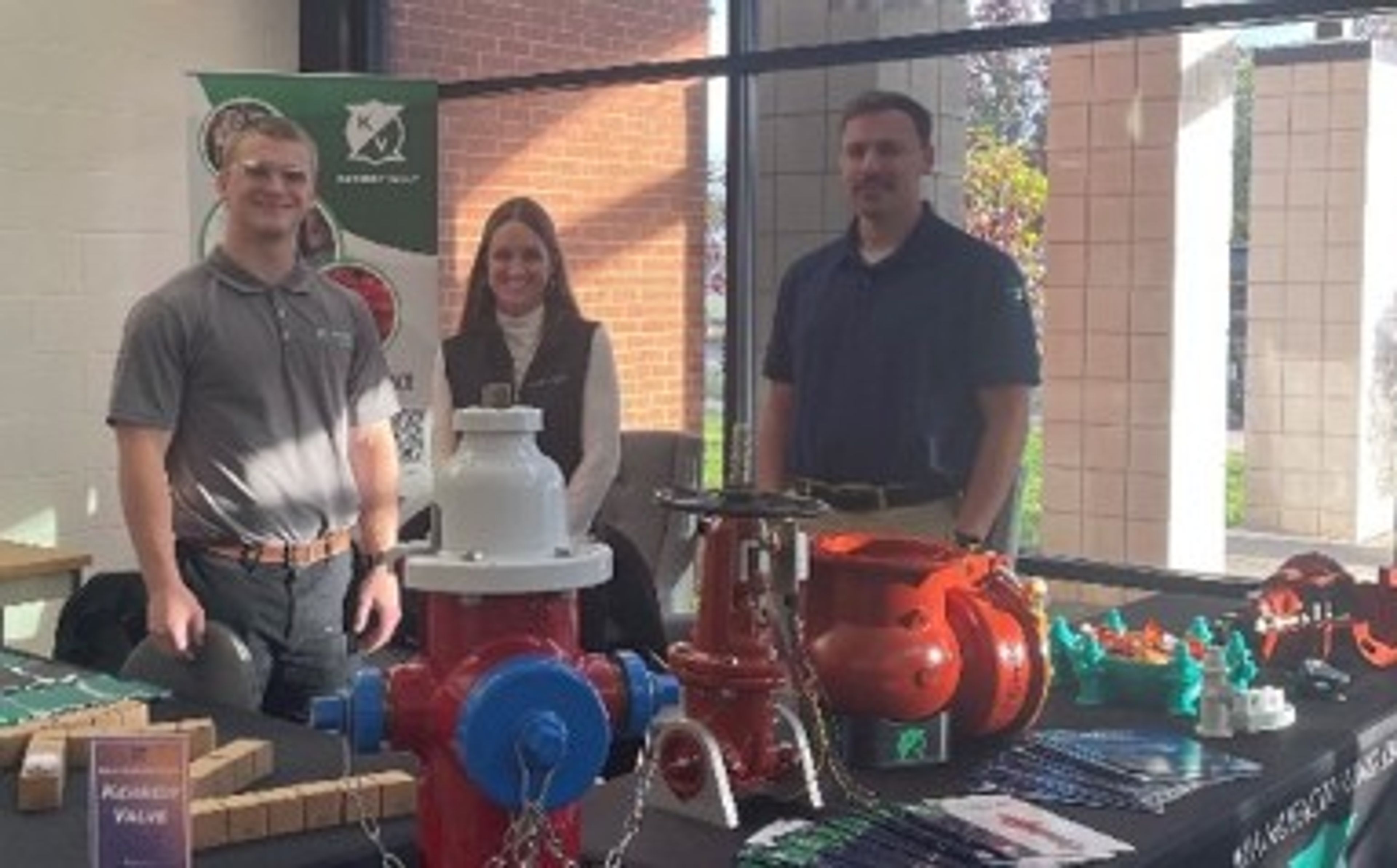 Three people stand behind a display table featuring industrial valves and piping equipment at an indoor event. Sunlight streams through large windows behind them, and promotional materials are arranged on the table.