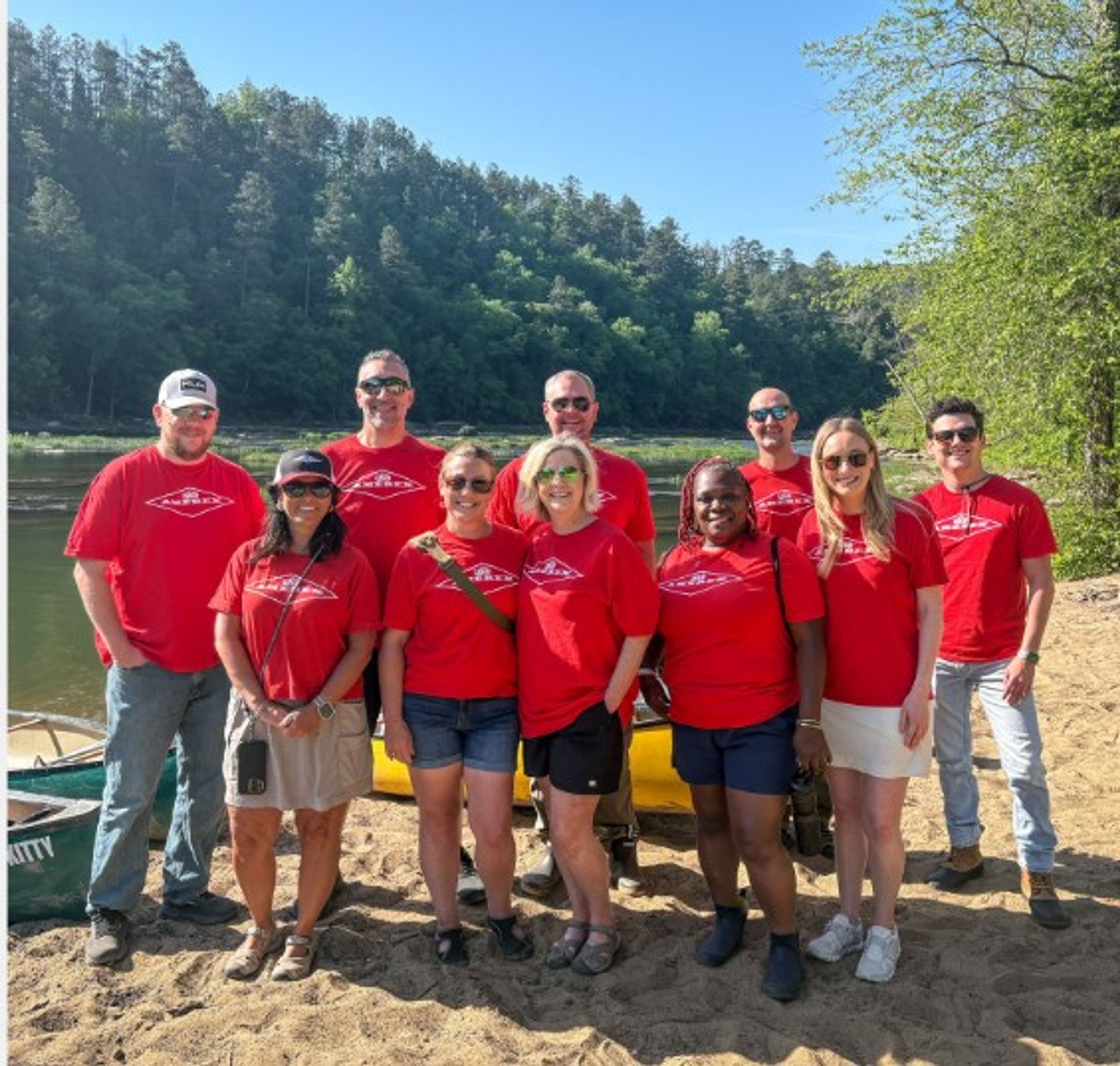 Ten people in matching red T-shirts pose together on a sandy riverbank with trees and a canoe in the background, smiling on a sunny day.