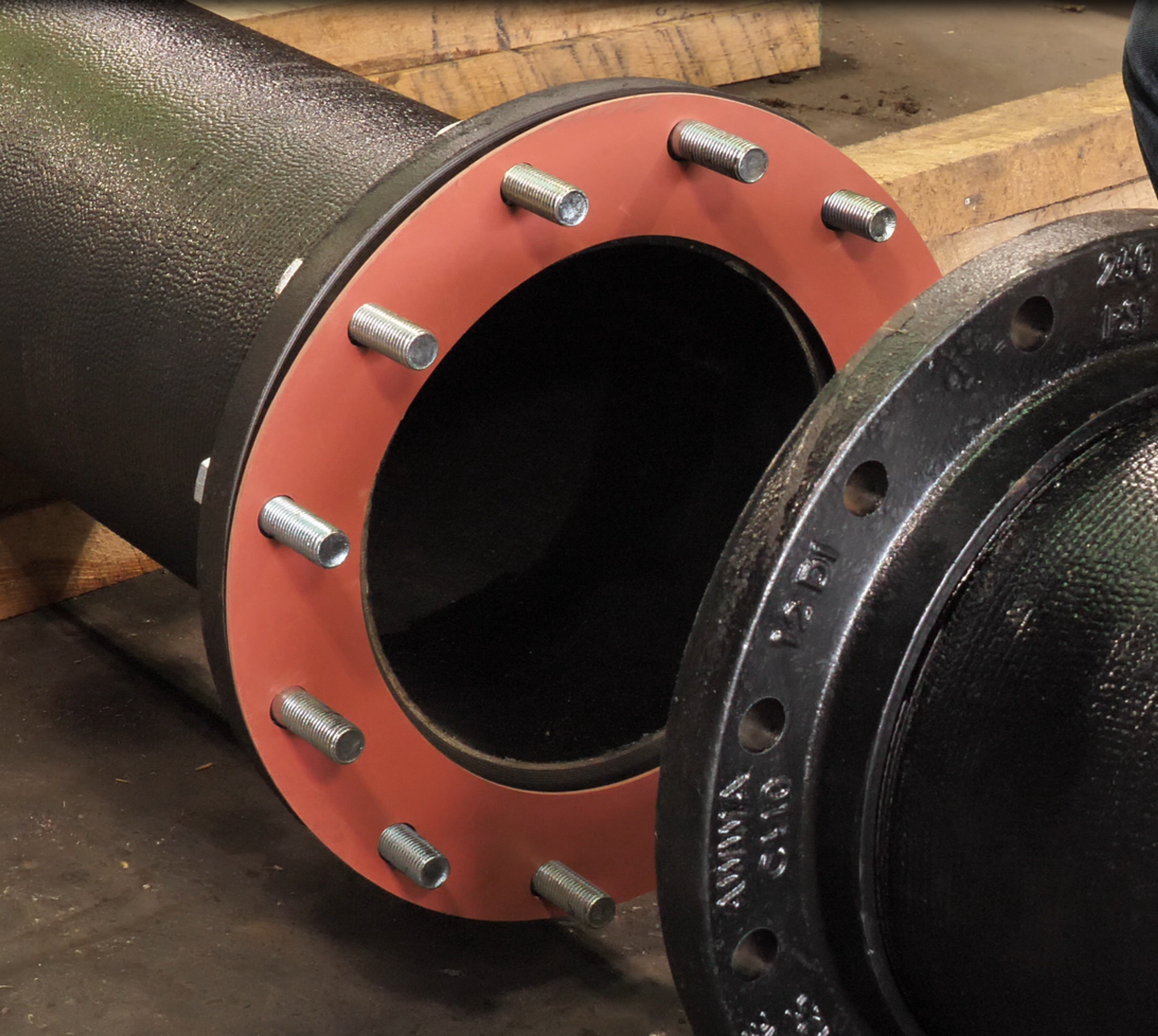 Two large industrial pipes with flanged ends are connected by bolts. The flange on the left has a red gasket between the pipe ends. Wooden supports are visible underneath the pipes.