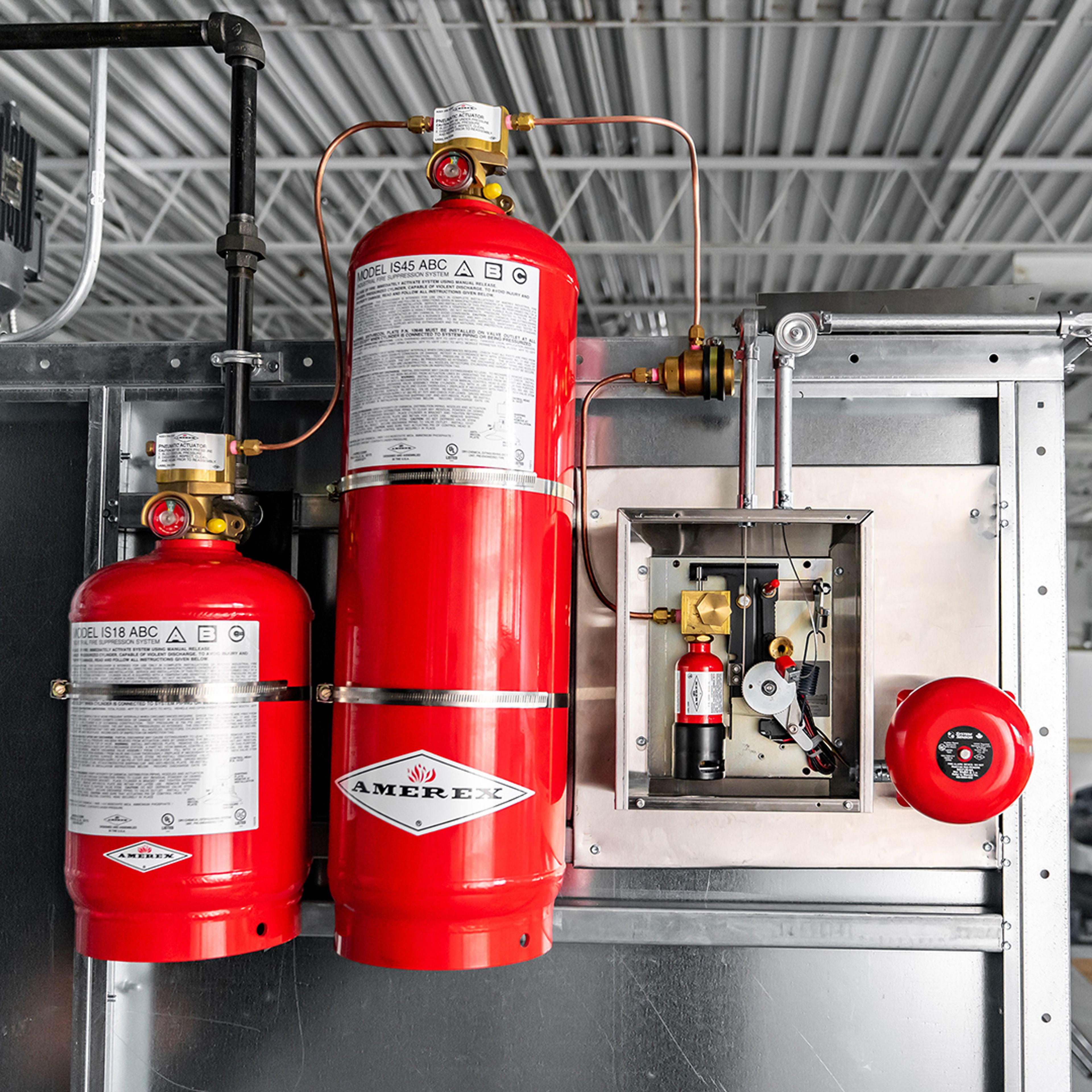 Two red Amerex fire suppression system tanks, control panel, and alarm bell mounted on a metal wall inside an industrial building with a gray exposed ceiling.