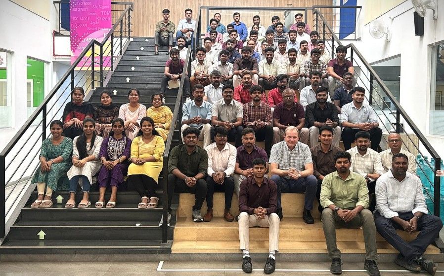A large group of people pose for a photo on indoor stadium-style seating, smiling and facing the camera. The setting appears to be a modern office or educational space.