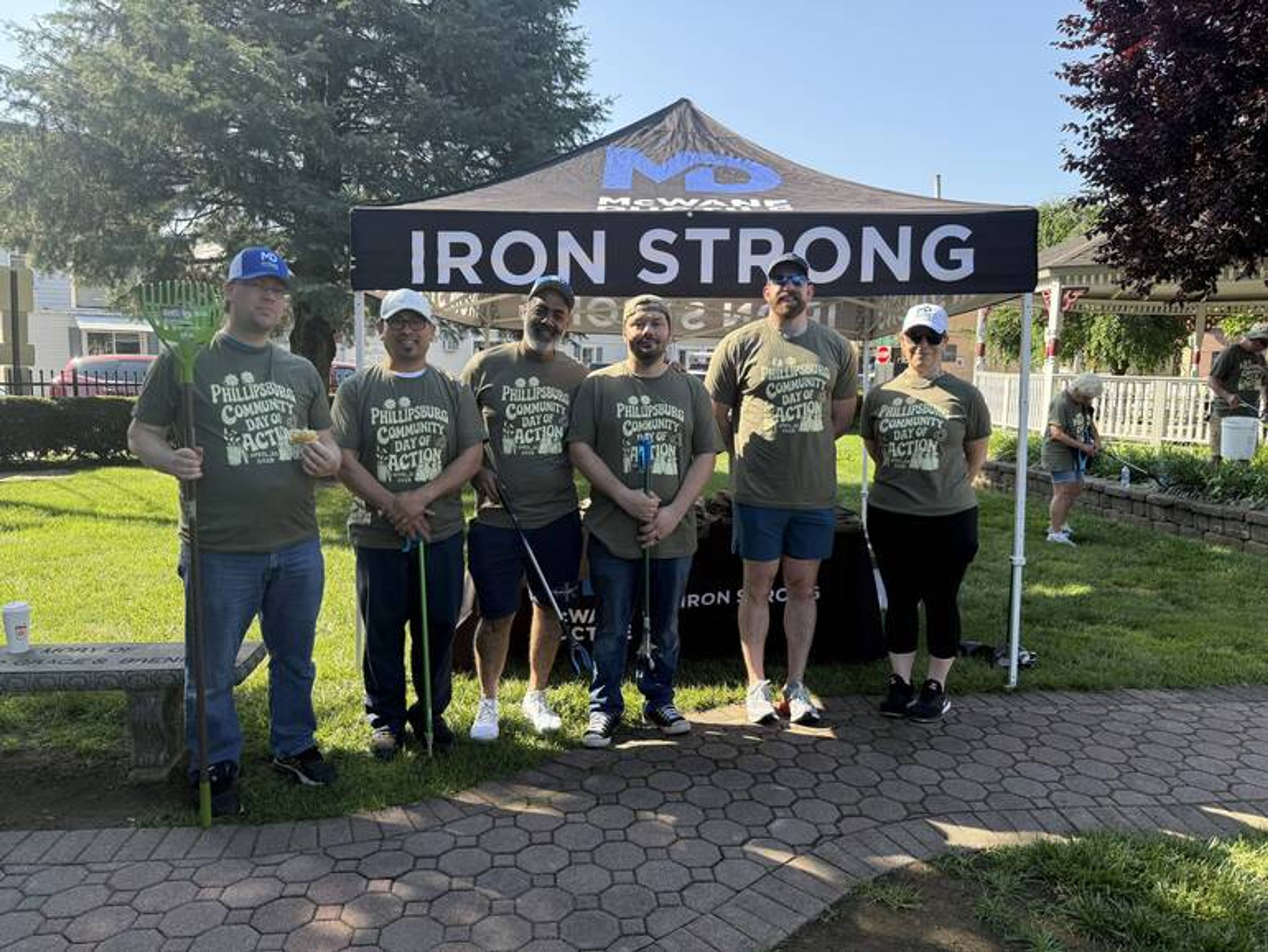 Six people stand in front of a black “IRON STRONG” canopy tent on grass, all wearing matching green T-shirts and holding trash grabbers, posing for a group photo at an outdoor park or community event.