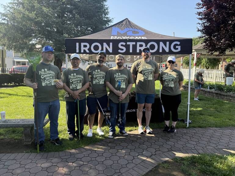 Six people stand in front of a black “IRON STRONG” canopy tent on grass, all wearing matching green T-shirts and holding trash grabbers, posing for a group photo at an outdoor park or community event.