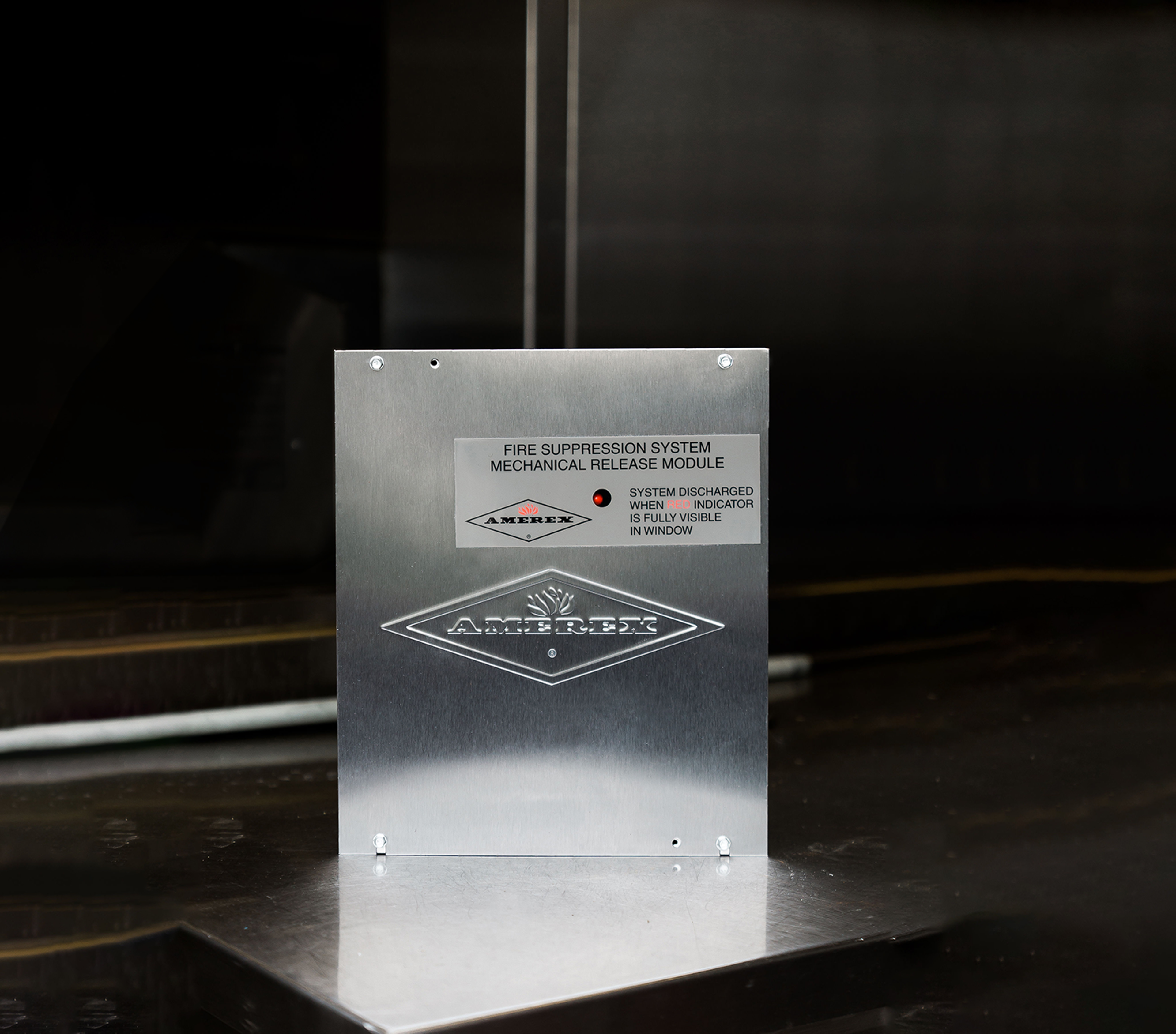 A metal box labeled “Fire Suppression System Mechanical Release Module” with the brand “Amerex” and instructions about the system’s status indicator, sits on a reflective metallic surface.