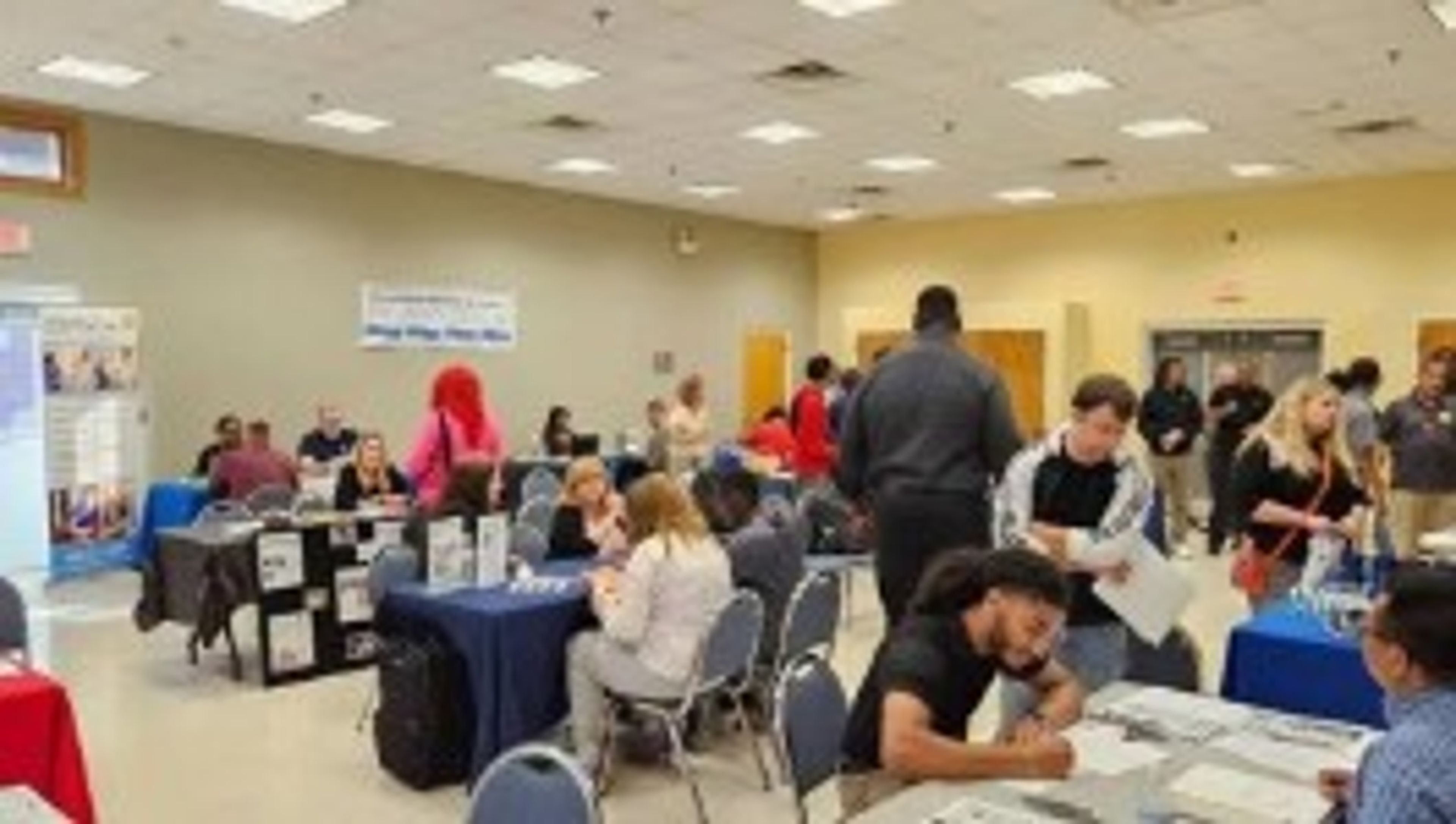 People attend a job fair in a large room with tables set up, staffed by recruiters. Attendees talk with representatives, fill out forms, and interact at various booths. The atmosphere is busy and engaged.