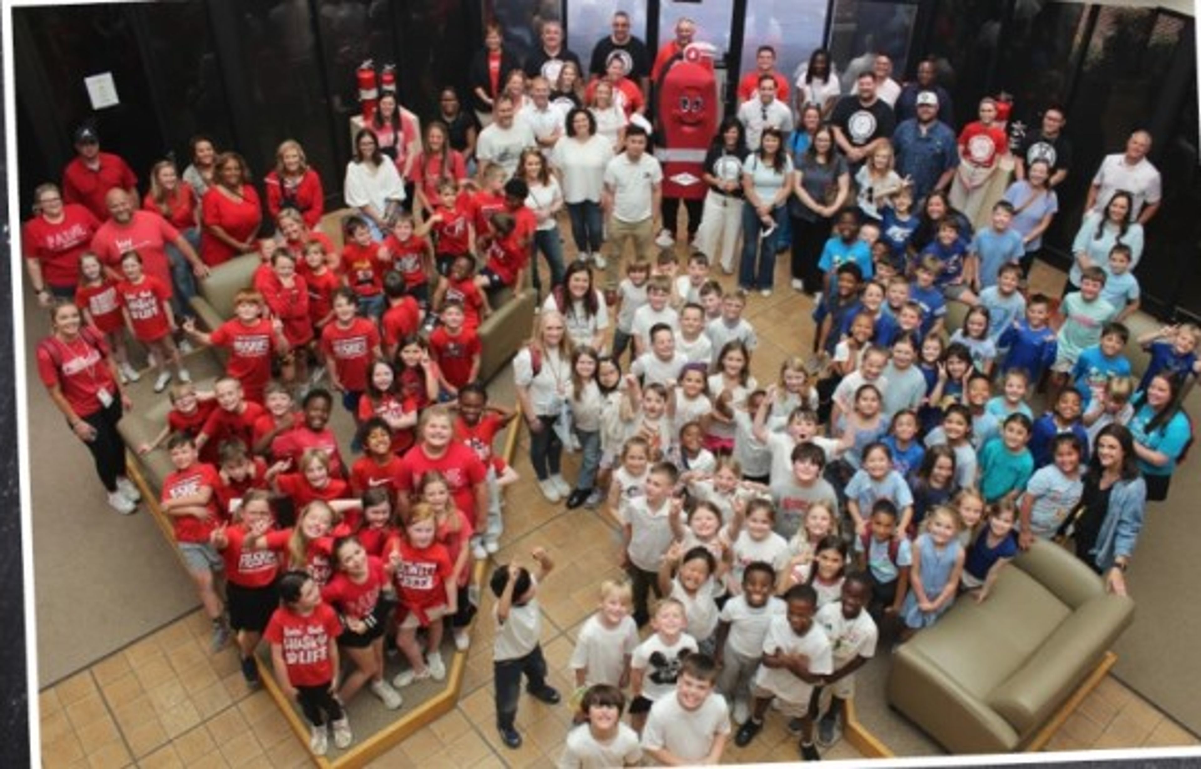 Large group of children and adults, some in matching shirts, pose together indoors around a mascot in a red costume, forming the shape of a heart. Some people are standing, while others are seated on couches.