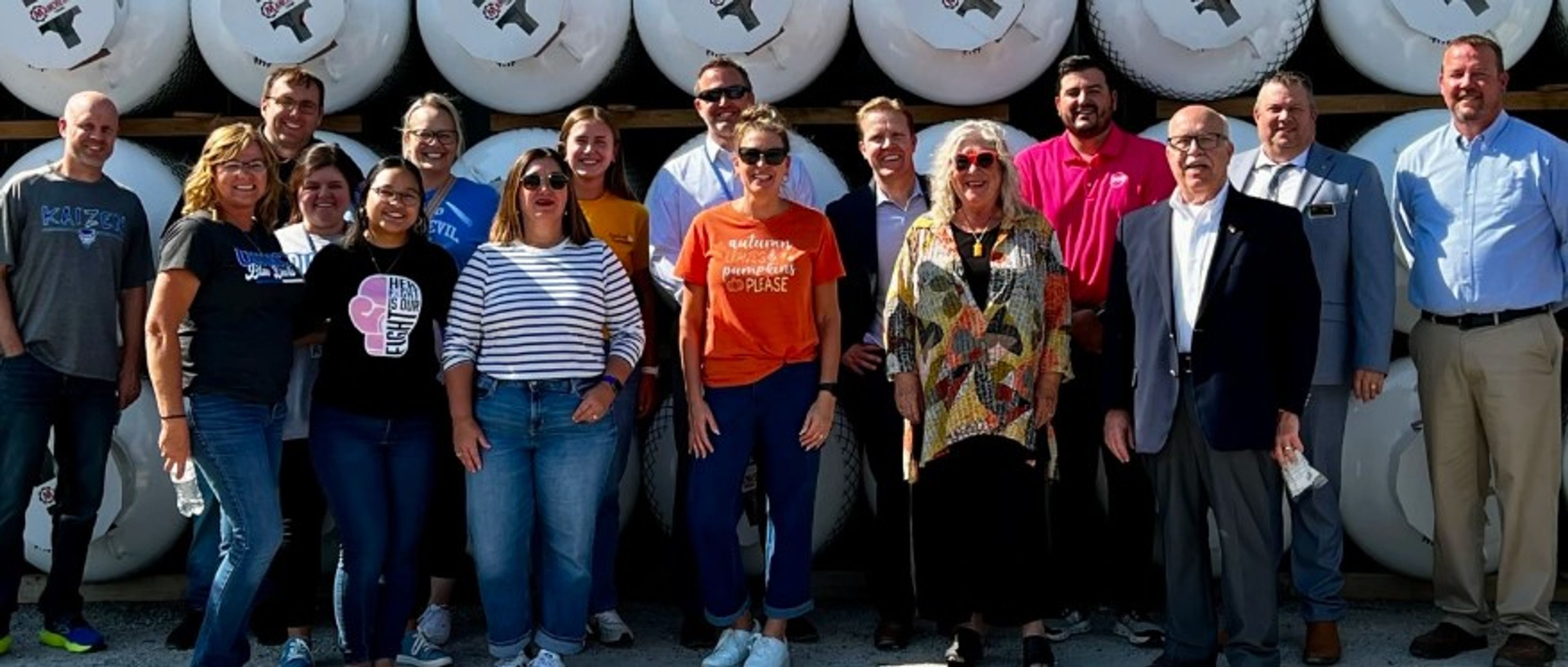 A group of fifteen adults stands smiling in front of stacked large white tanks outdoors. Most are casually dressed; a few wear business attire. It is sunny and everyone is posing closely together for the photo.
