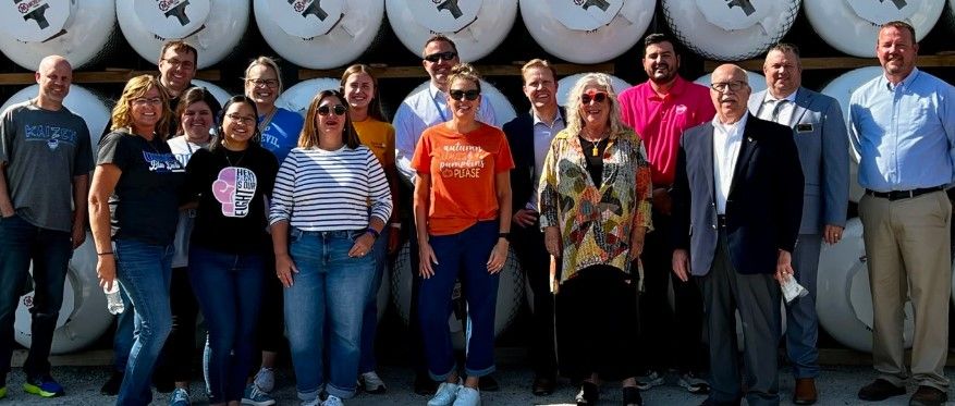 A group of fifteen adults stands smiling in front of stacked large white tanks outdoors. Most are casually dressed; a few wear business attire. It is sunny and everyone is posing closely together for the photo.