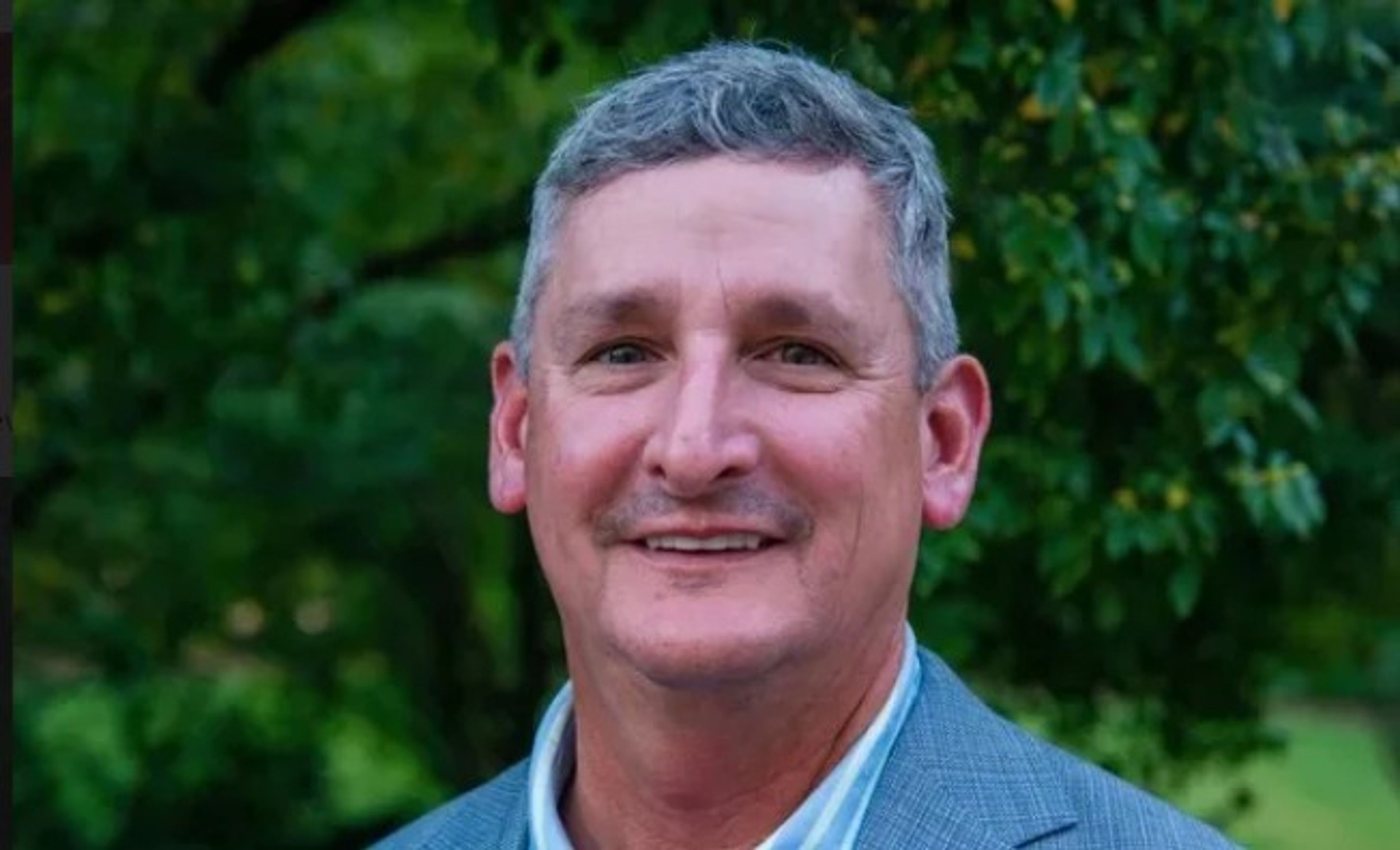 A middle-aged man with short gray hair and a mustache, wearing a gray suit jacket and striped shirt, smiles outdoors with trees and greenery in the background.