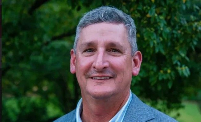 A middle-aged man with short gray hair and a mustache, wearing a gray suit jacket and striped shirt, smiles outdoors with trees and greenery in the background.