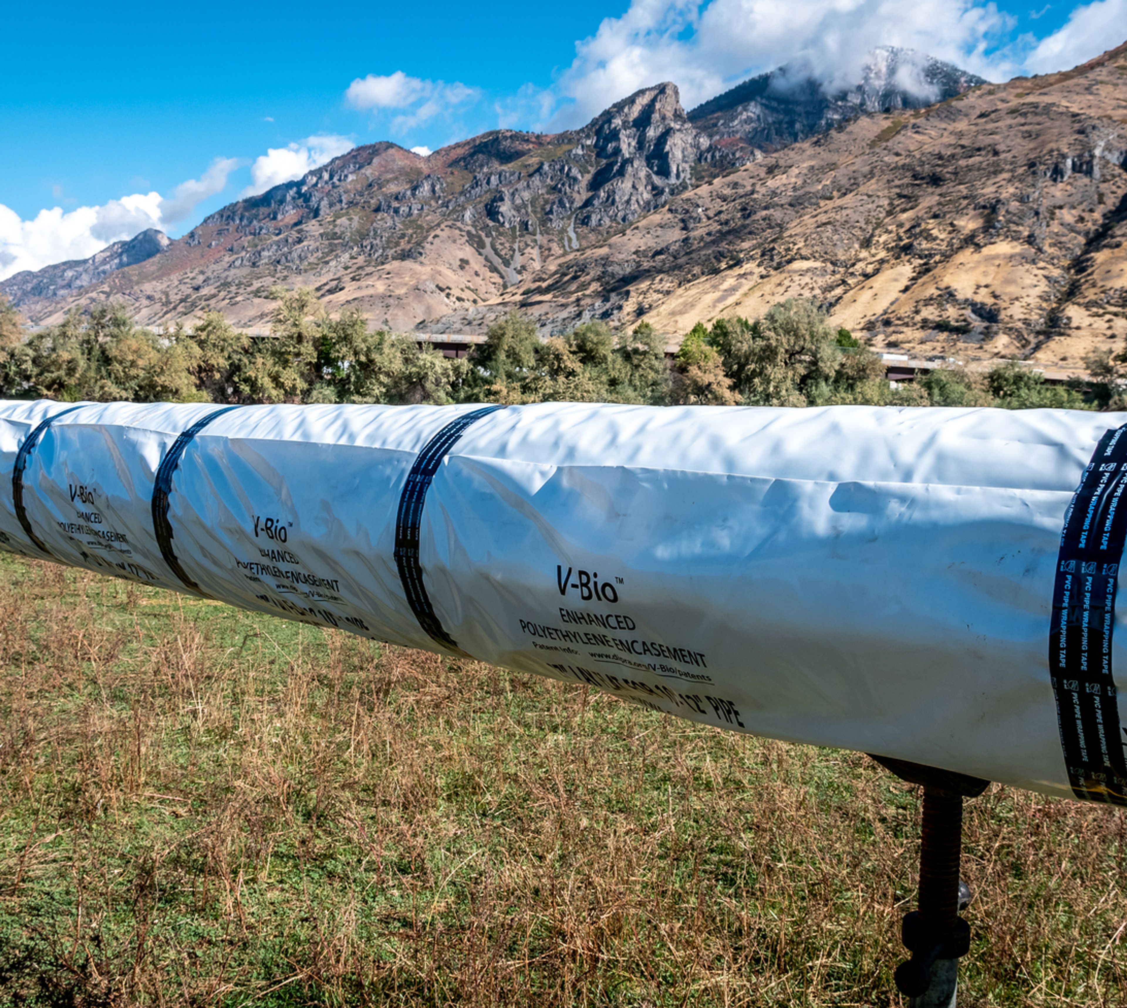 A large, white plastic-wrapped pipe labeled “V-Bio” runs through a grassy field with mountains and trees in the background under a partly cloudy sky.