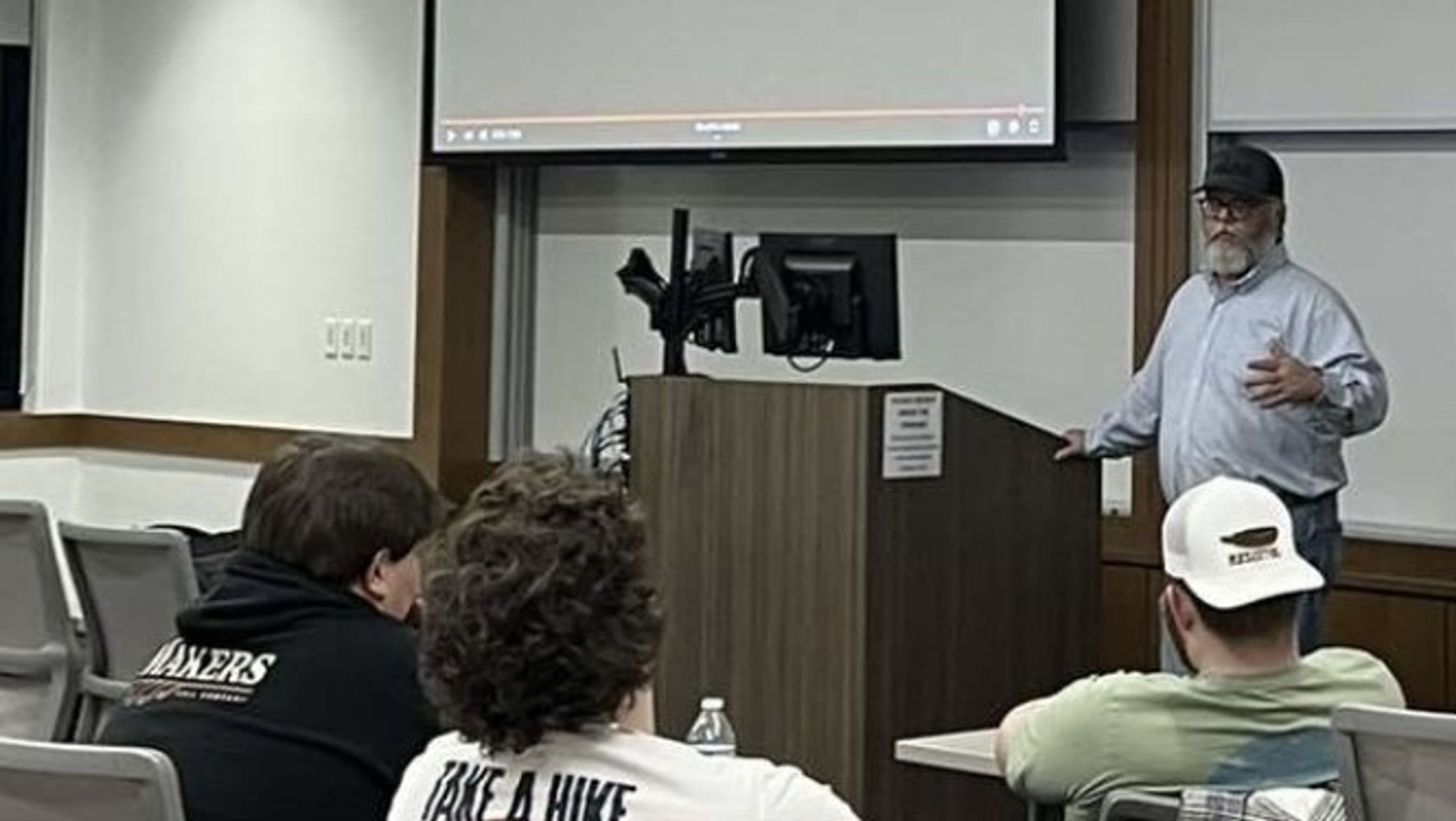 A man with a beard and cap stands at a podium giving a lecture to several seated students in a classroom with a large screen behind him.