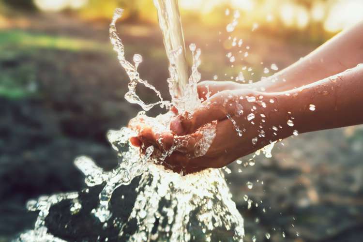 Hands cupping clear, flowing water as it splashes in sunlight, with a blurred natural background.