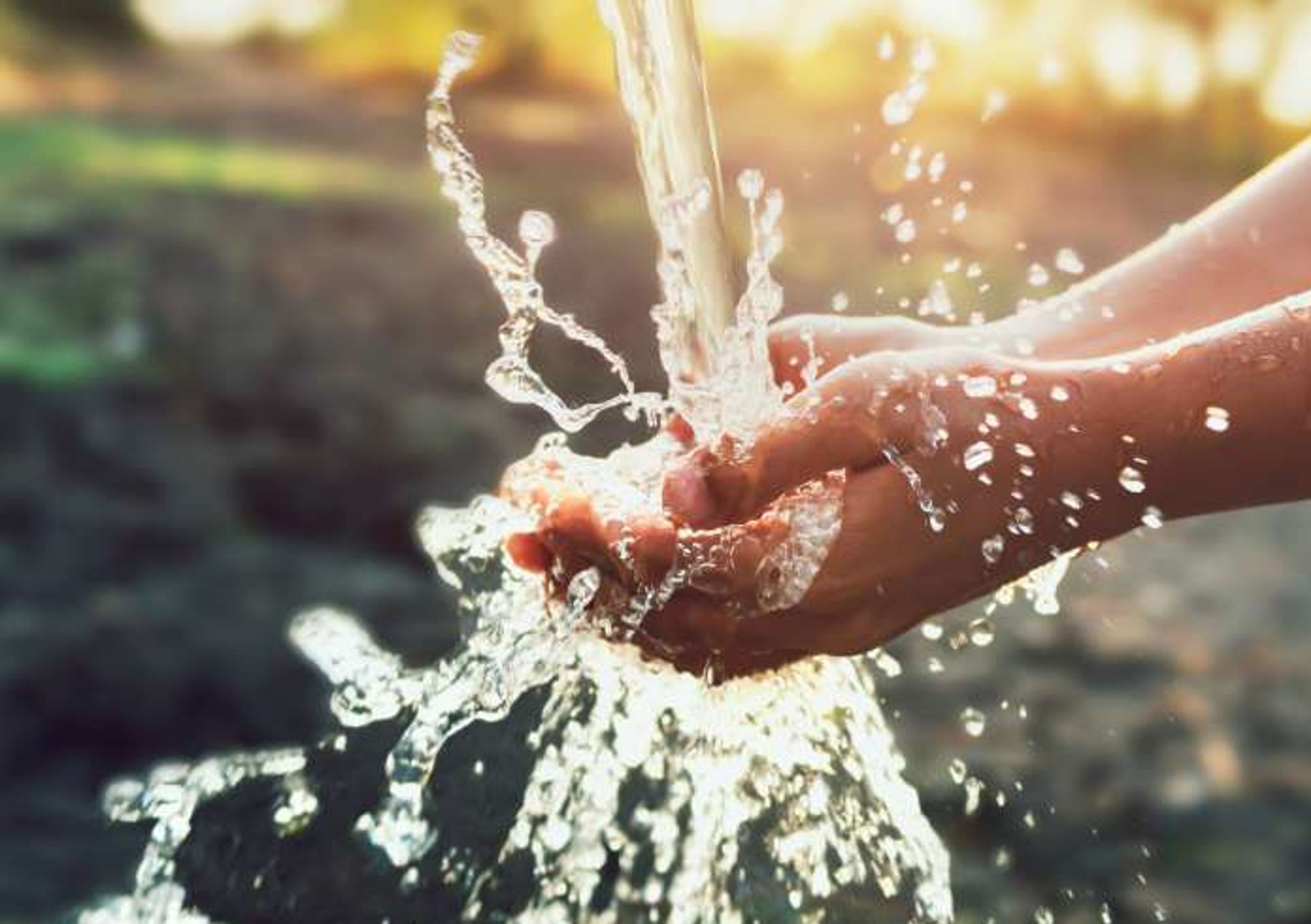 Hands cupping clear, flowing water as it splashes in sunlight, with a blurred natural background.