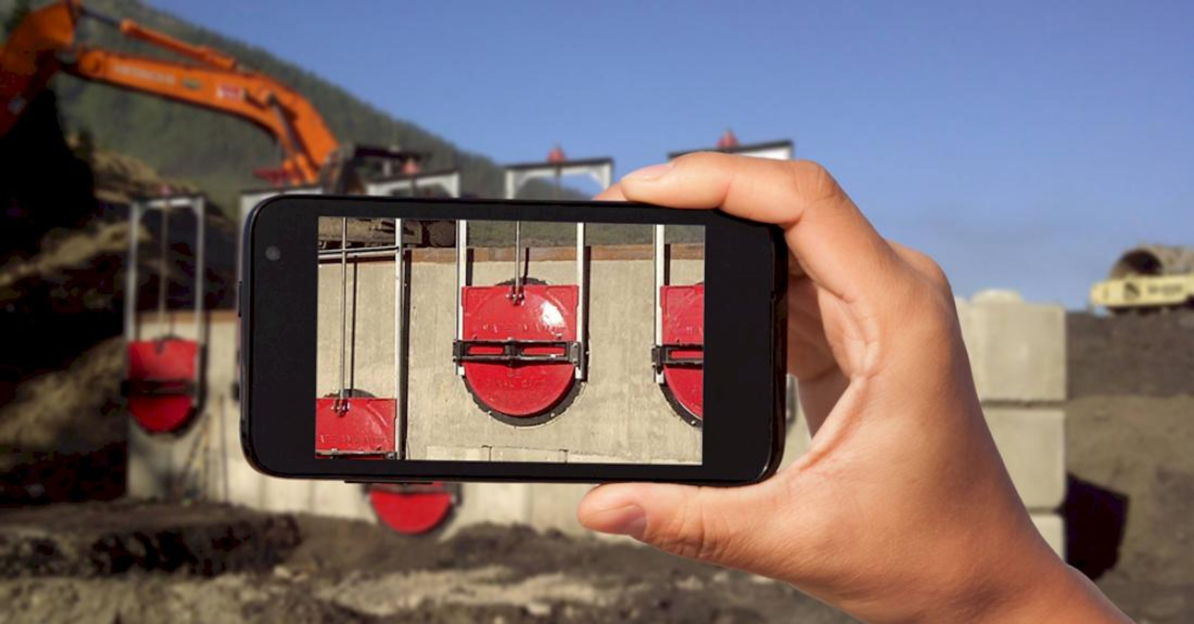 A hand holds a smartphone, taking a photo of red circular valves on a concrete wall at a construction site with an excavator and dirt in the background.
