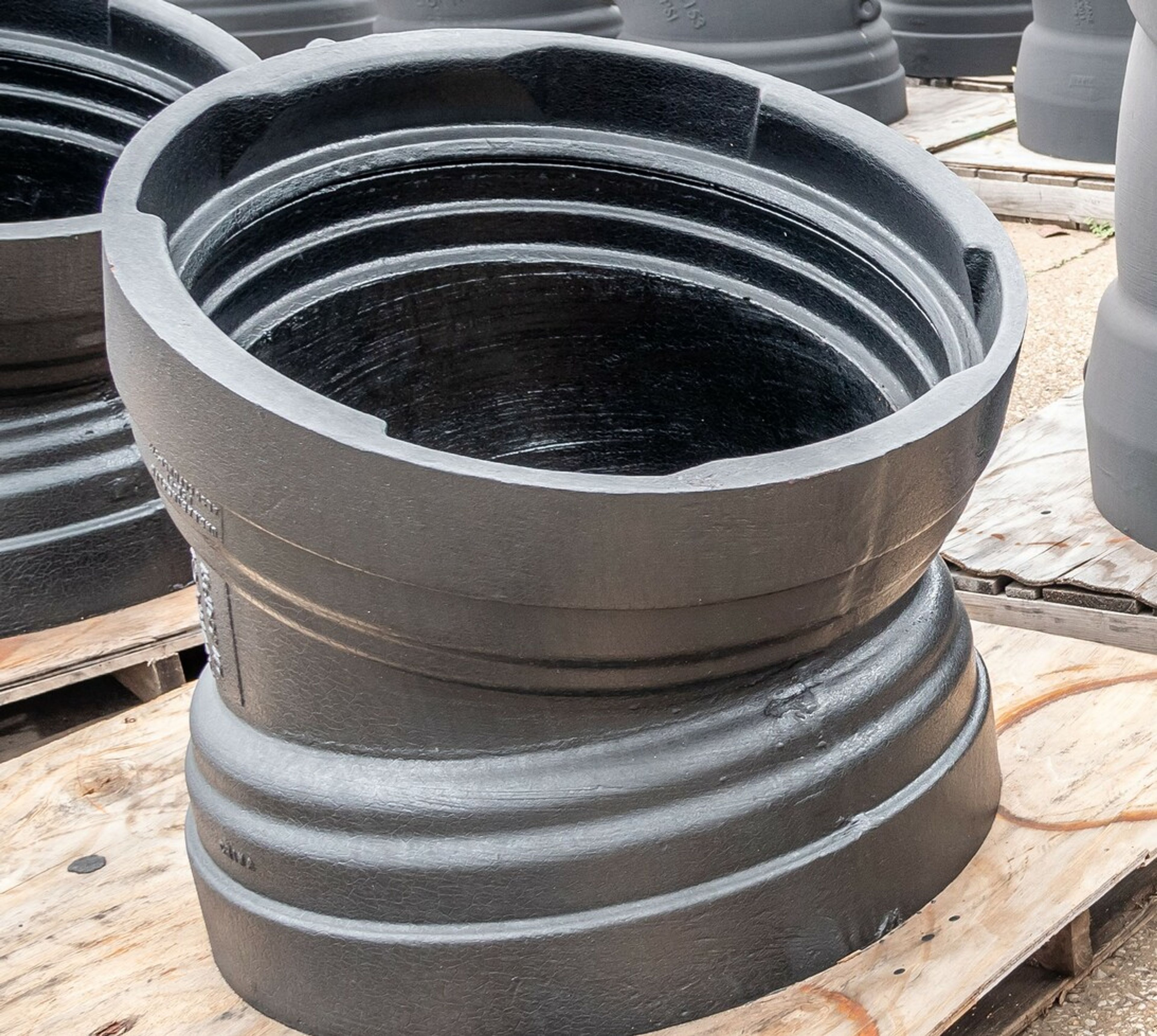 A large, black, industrial metal pipe fitting with a slanted opening sits on a wooden pallet outdoors, surrounded by similar pipe fittings.