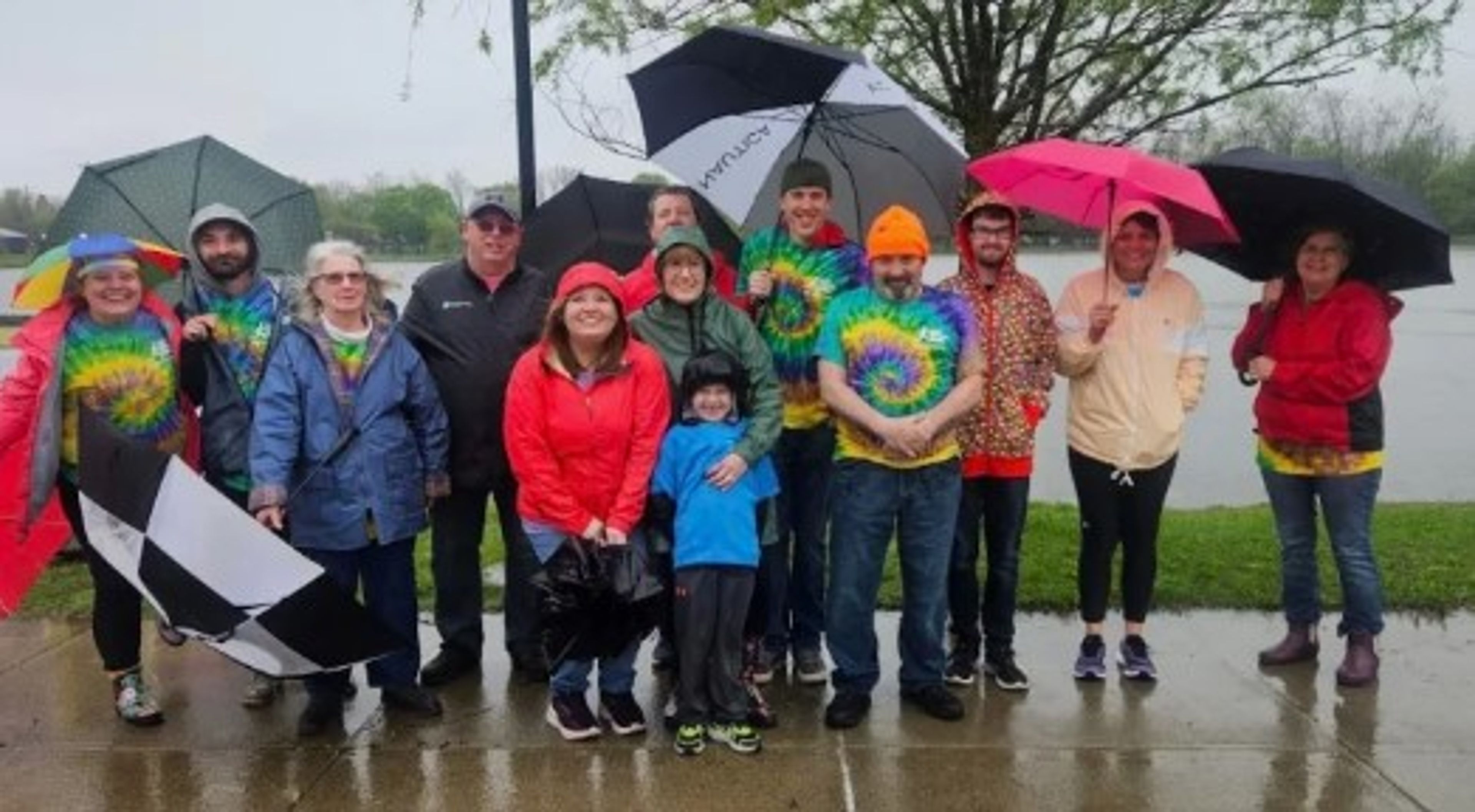A group of people in colorful rain gear and tie-dye shirts stand together on a rainy day, holding umbrellas and smiling at the camera near a body of water.