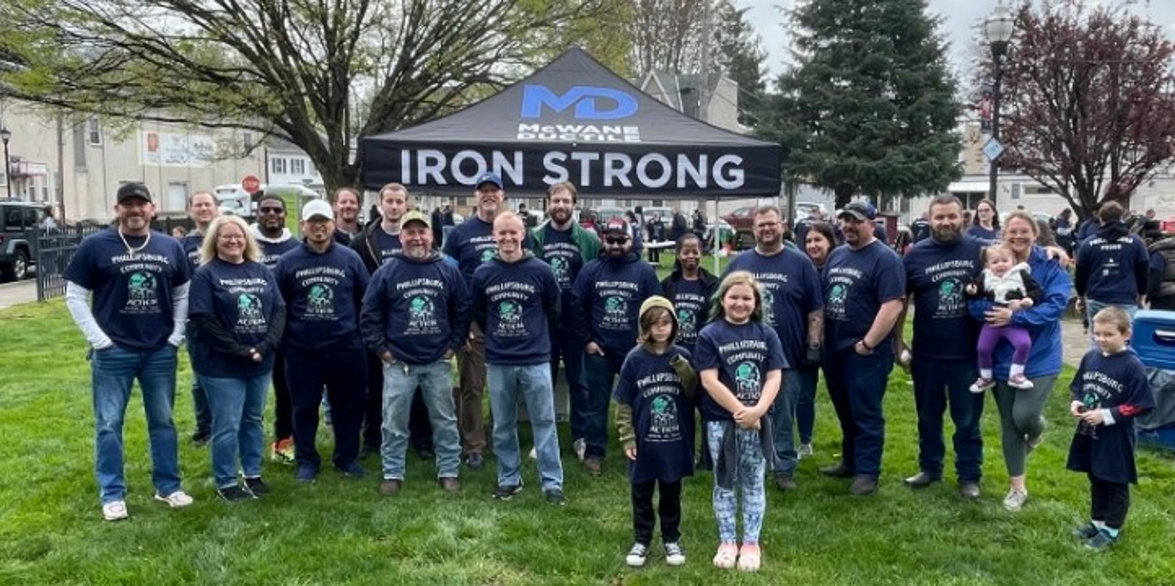 A group of people, including children, pose in front of a tent labeled “IRON STRONG” on a grassy area. Most are wearing matching dark shirts. Trees and buildings are visible in the background.
