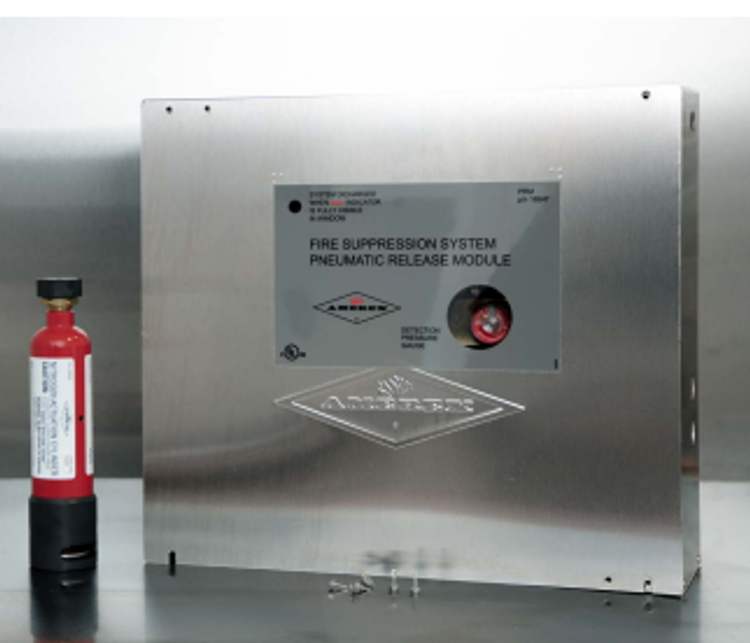 A metal fire suppression control panel labeled “Pneumatic Release Module” is mounted on a wall. A small red fire suppressant canister stands upright next to it on a metallic surface.