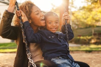 A smiling woman and child sit together on a swing at a playground, enjoying the sunshine.