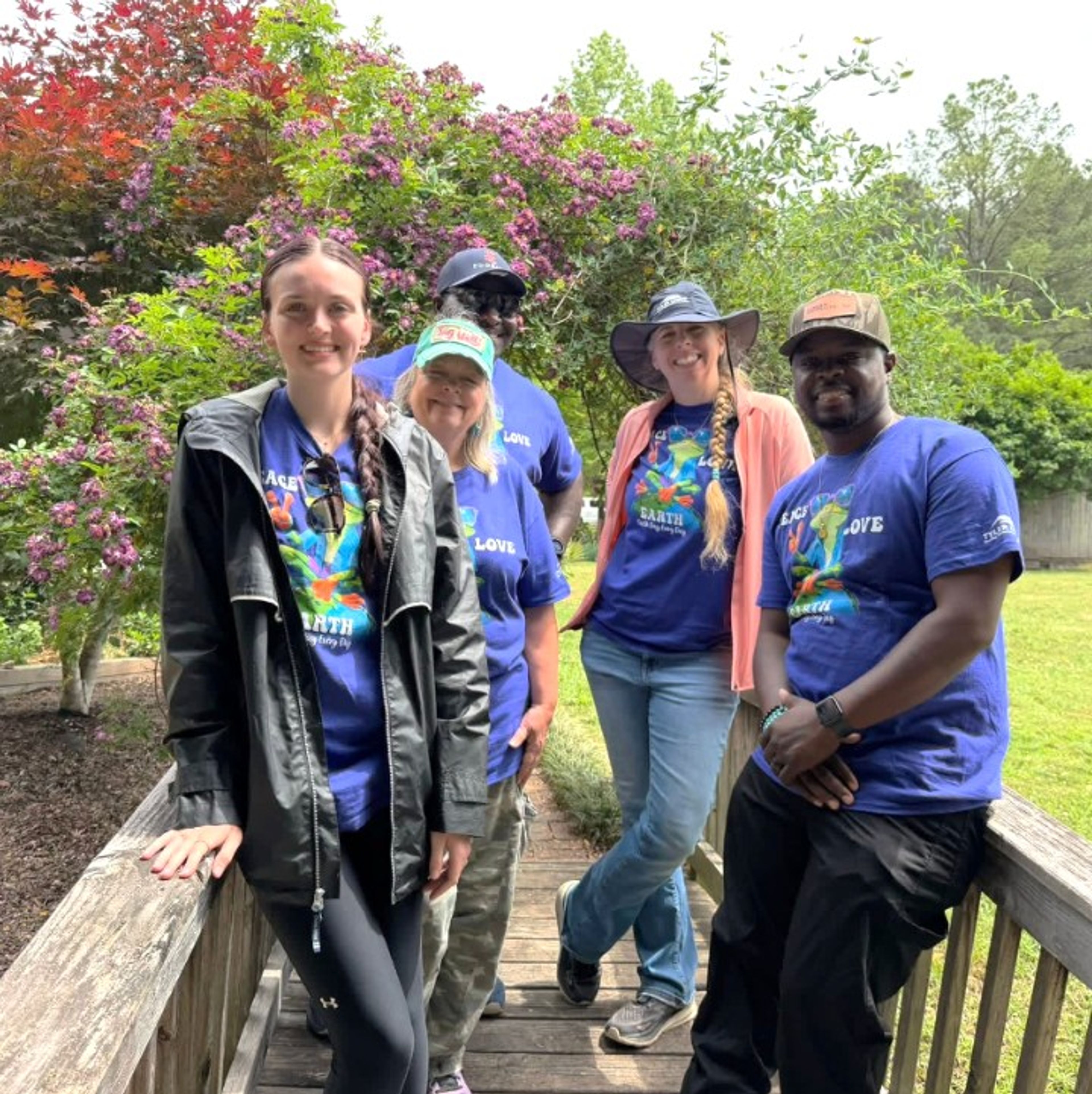 Five people stand smiling on a wooden bridge in a garden, wearing blue Earth Day shirts and hats. Green grass and blooming trees are visible in the background.