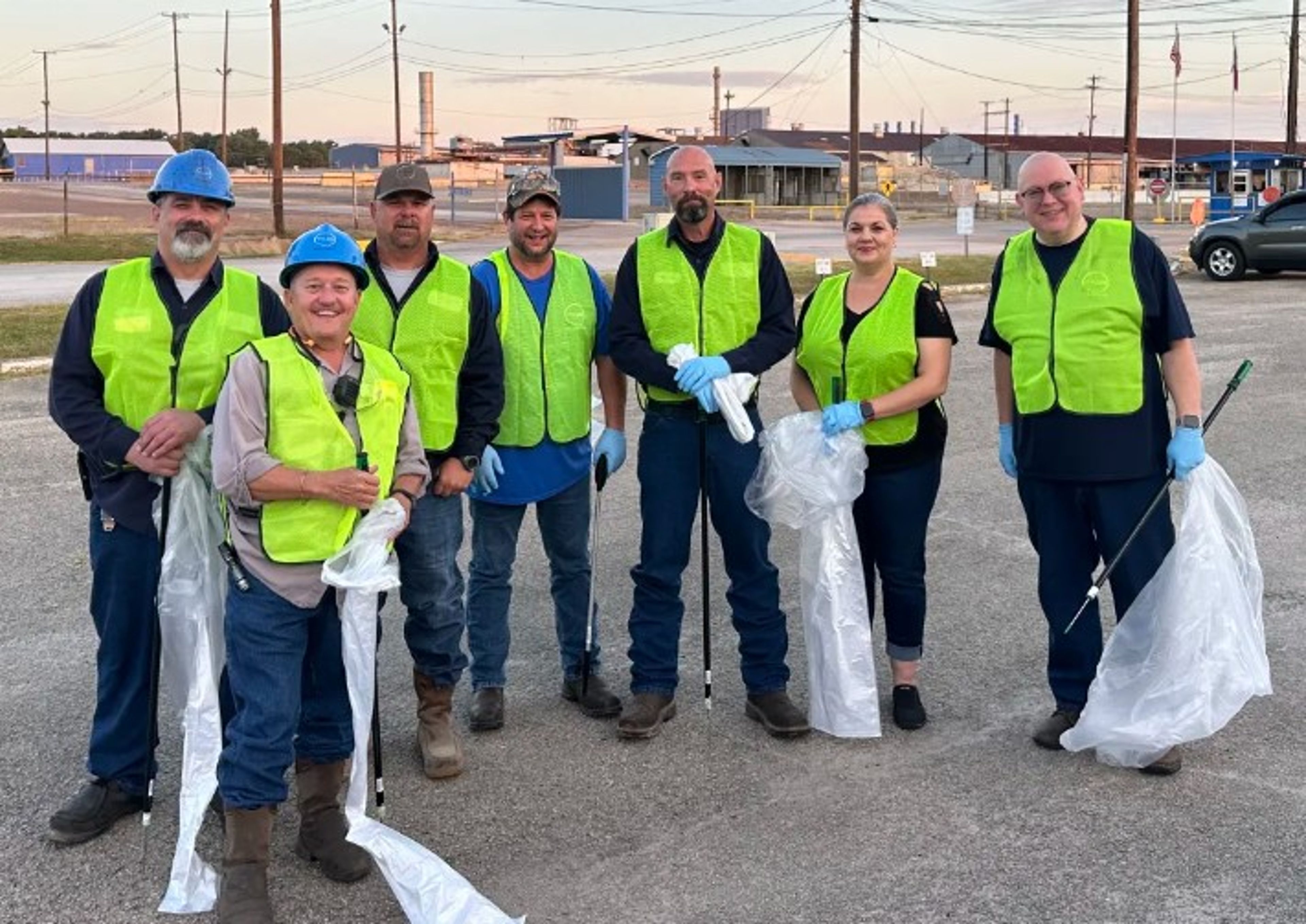 Seven people wearing yellow safety vests, gloves, and hard hats stand in a parking lot holding trash grabbers and bags, posing for a group photo during a cleanup event. Industrial buildings are visible in the background.