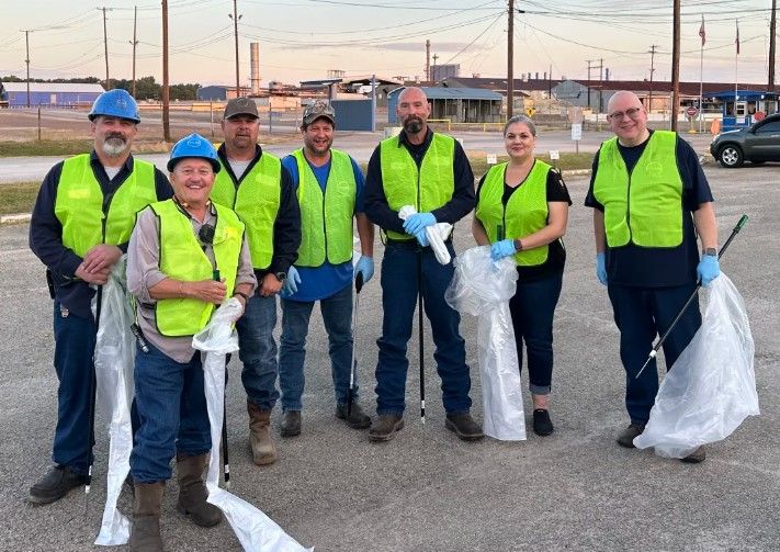 Seven people wearing yellow safety vests, gloves, and hard hats stand in a parking lot holding trash grabbers and bags, posing for a group photo during a cleanup event. Industrial buildings are visible in the background.
