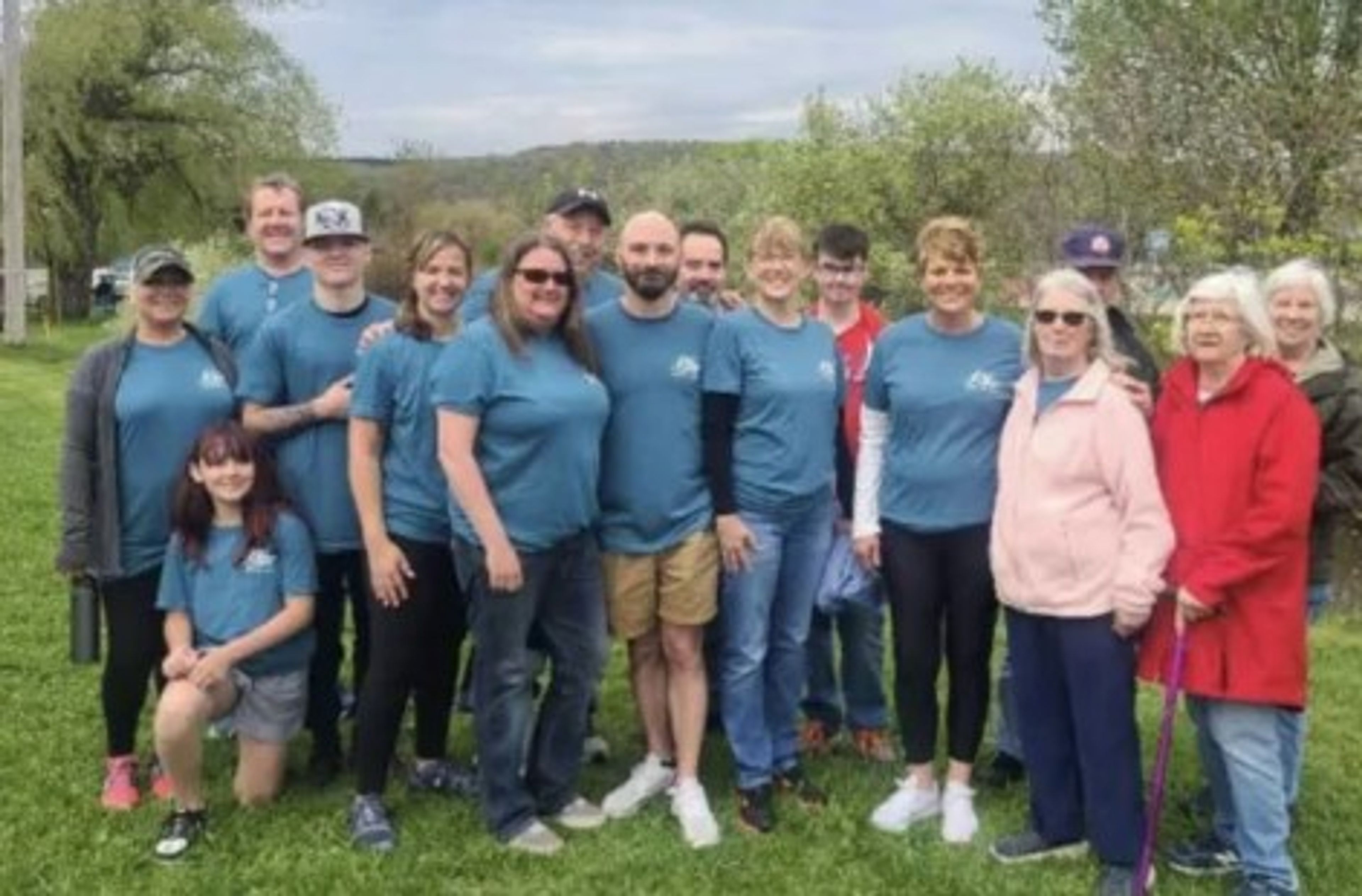A group of people, many wearing matching blue shirts, stand and smile outdoors on grass with trees in the background.