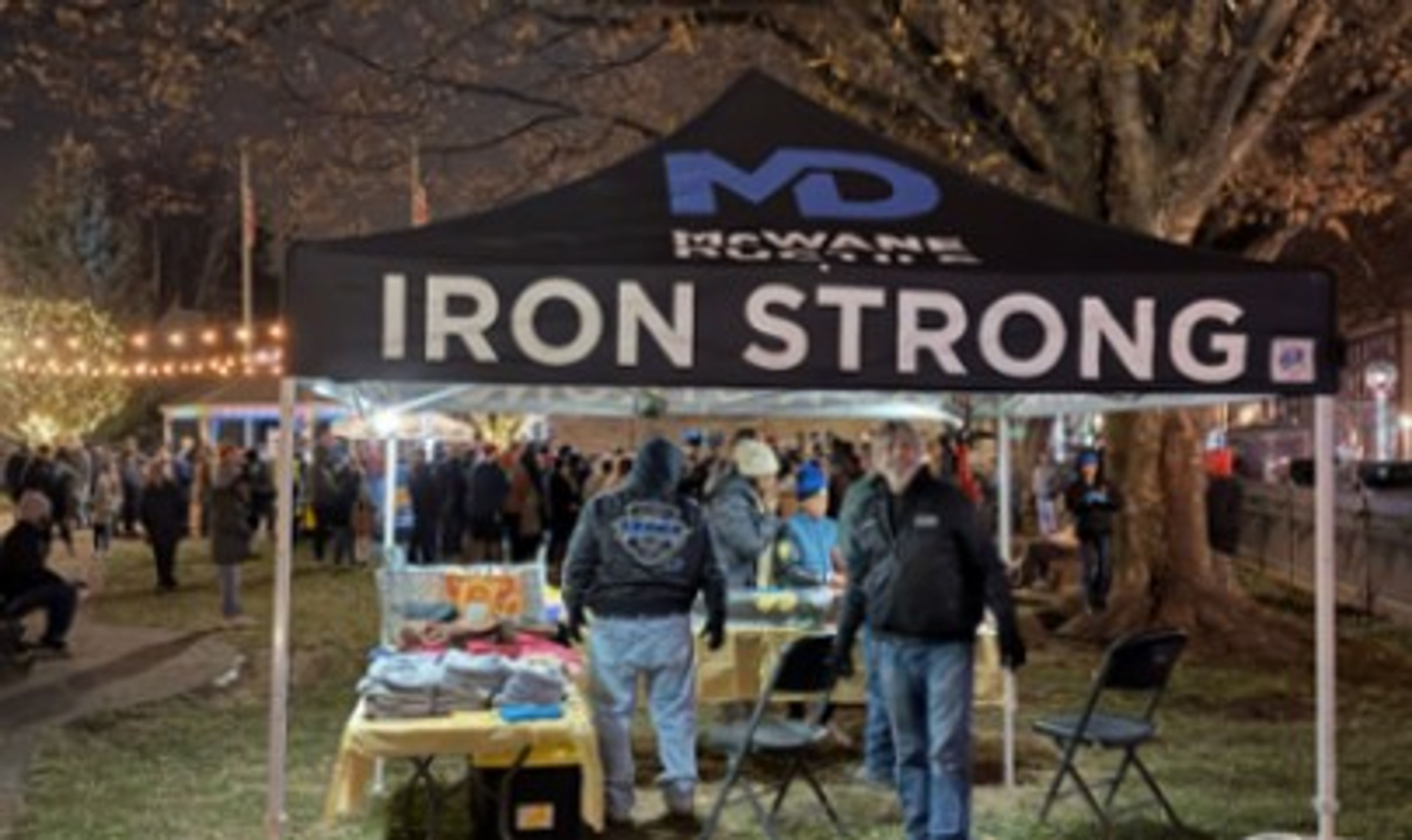 A black tent with “IRON STRONG” written on it stands at an outdoor night event. People gather under the tent and in the background, with string lights and trees visible.