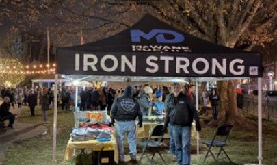 A black tent with “IRON STRONG” written on it stands at an outdoor night event. People gather under the tent and in the background, with string lights and trees visible.